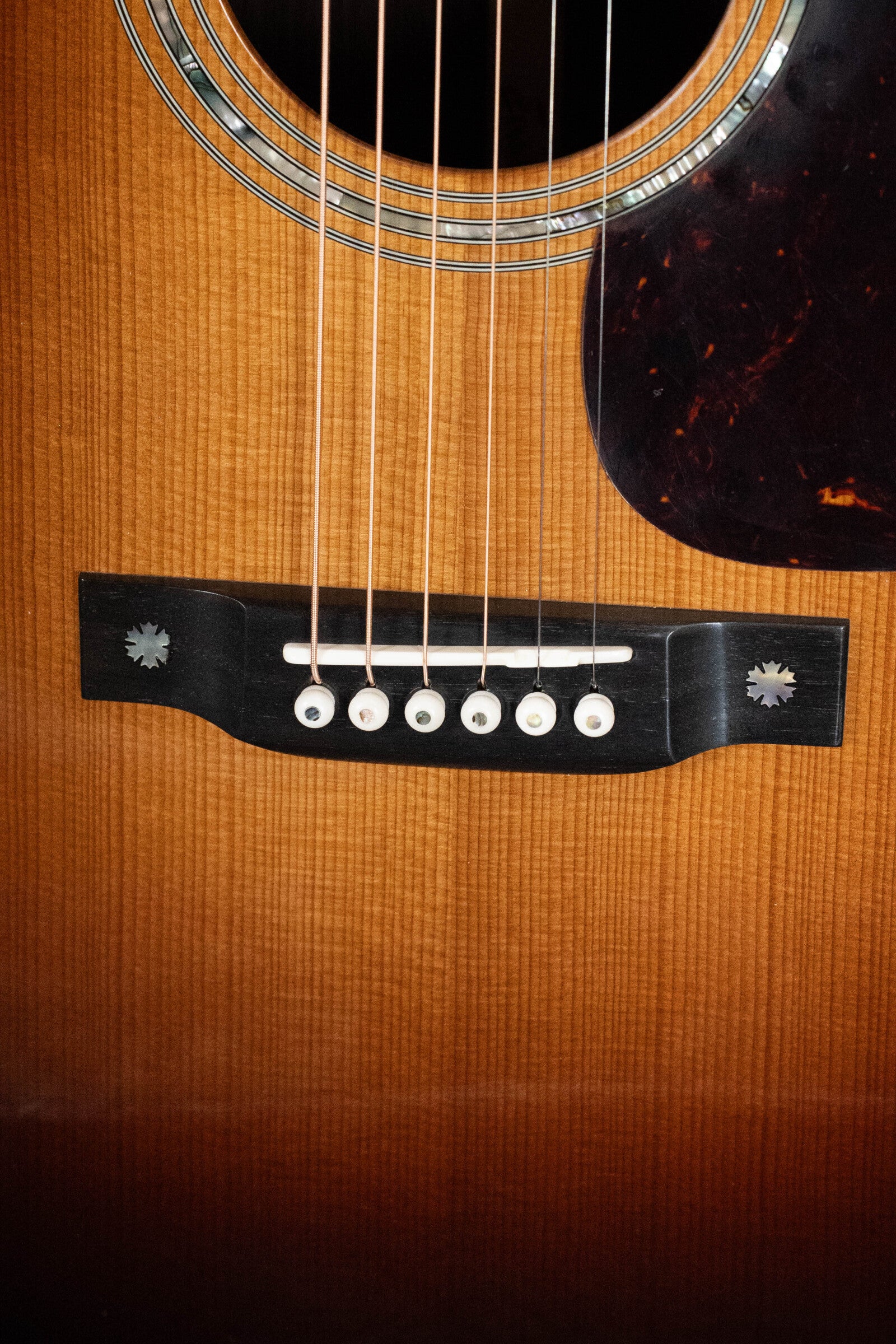 Close-up of the Eastman Guitars E40D-TC-SB Sunburst Dreadnought #1113 shows six strings, a wooden bridge with white pins, plus part of the sound hole and pickguard. Features thermo-cured AAA Adirondack top and rosewood back and sides.