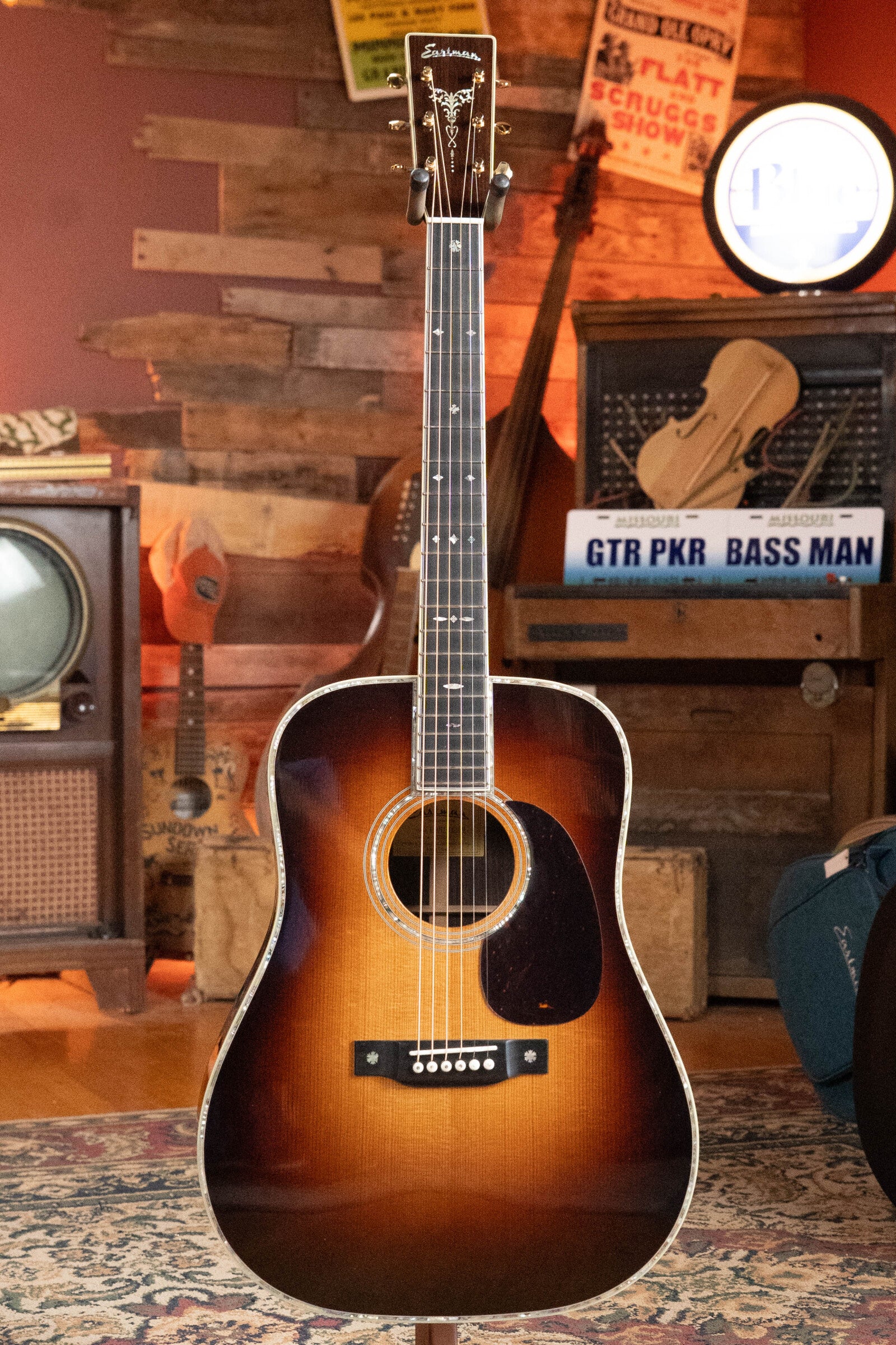 An Eastman Guitars E40D-TC-SB Thermo-cured AAA Adirondack/Rosewood Sunburst Dreadnought Acoustic Guitar #1113 stands on a patterned rug in a cozy room with vintage wooden decor, old radios, and music-themed signs.