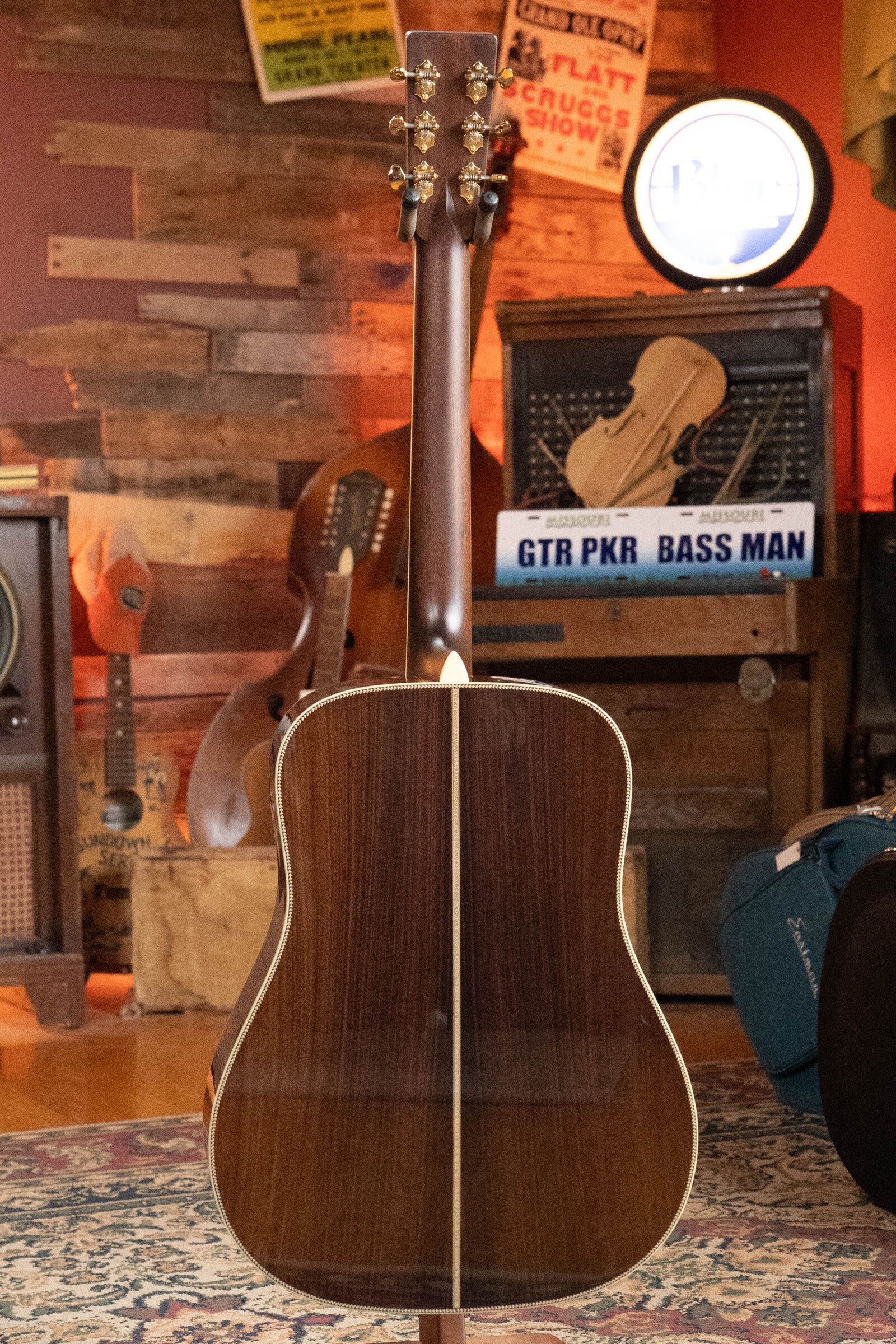 An Eastman Guitars E40D-TC-SB Thermo-cured AAA Grade Adirondack/Rosewood Sunburst Dreadnought Acoustic Guitar #1113 stands upright, back facing the camera in a cozy room with vintage instruments, wood panels, and music-themed signs.