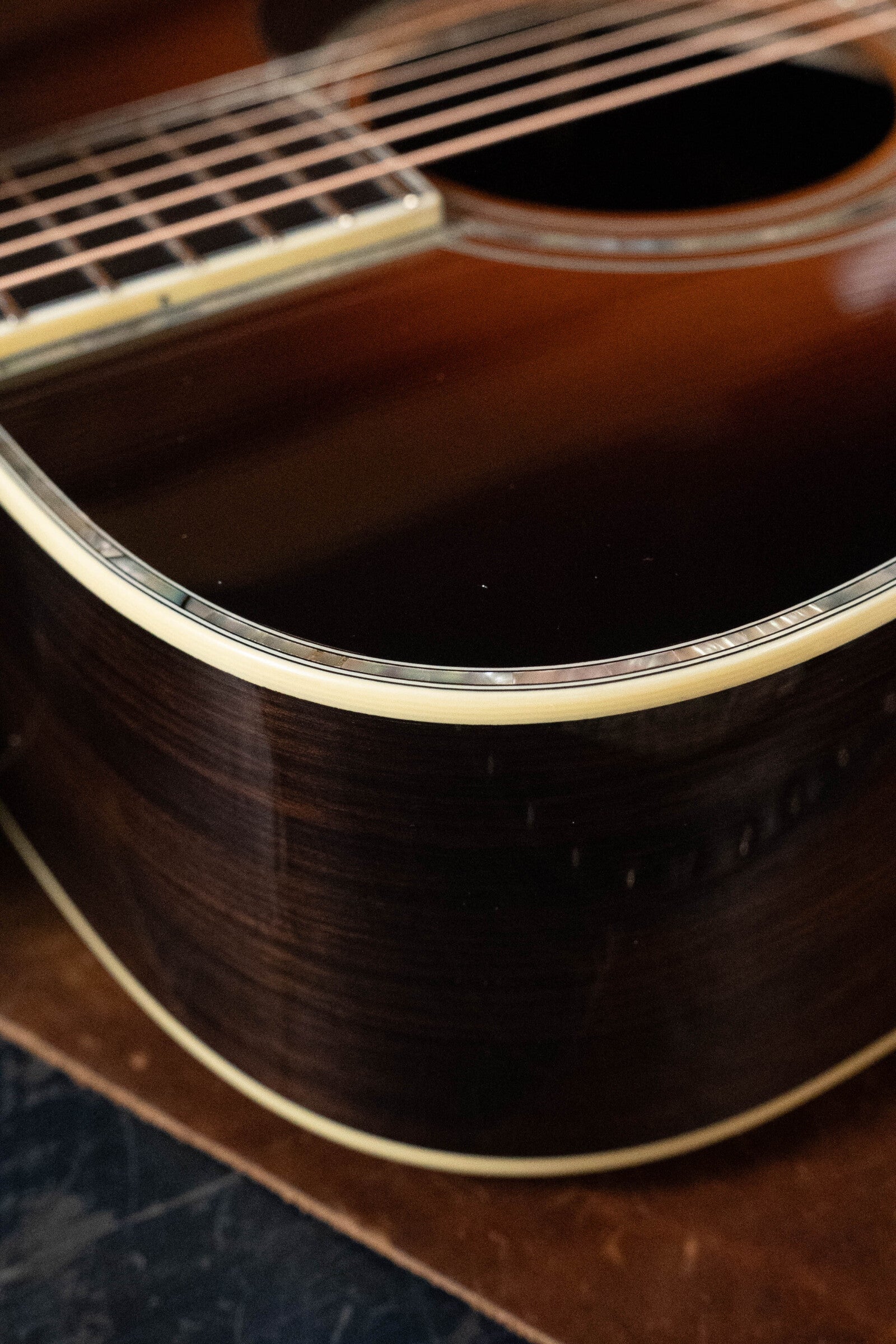 Close-up of an Eastman Guitars E40D-TC-SB #1113 Dreadnought showing the glossy rosewood back and sides, cream binding, and part of the fretboard, resting on a brown surface.