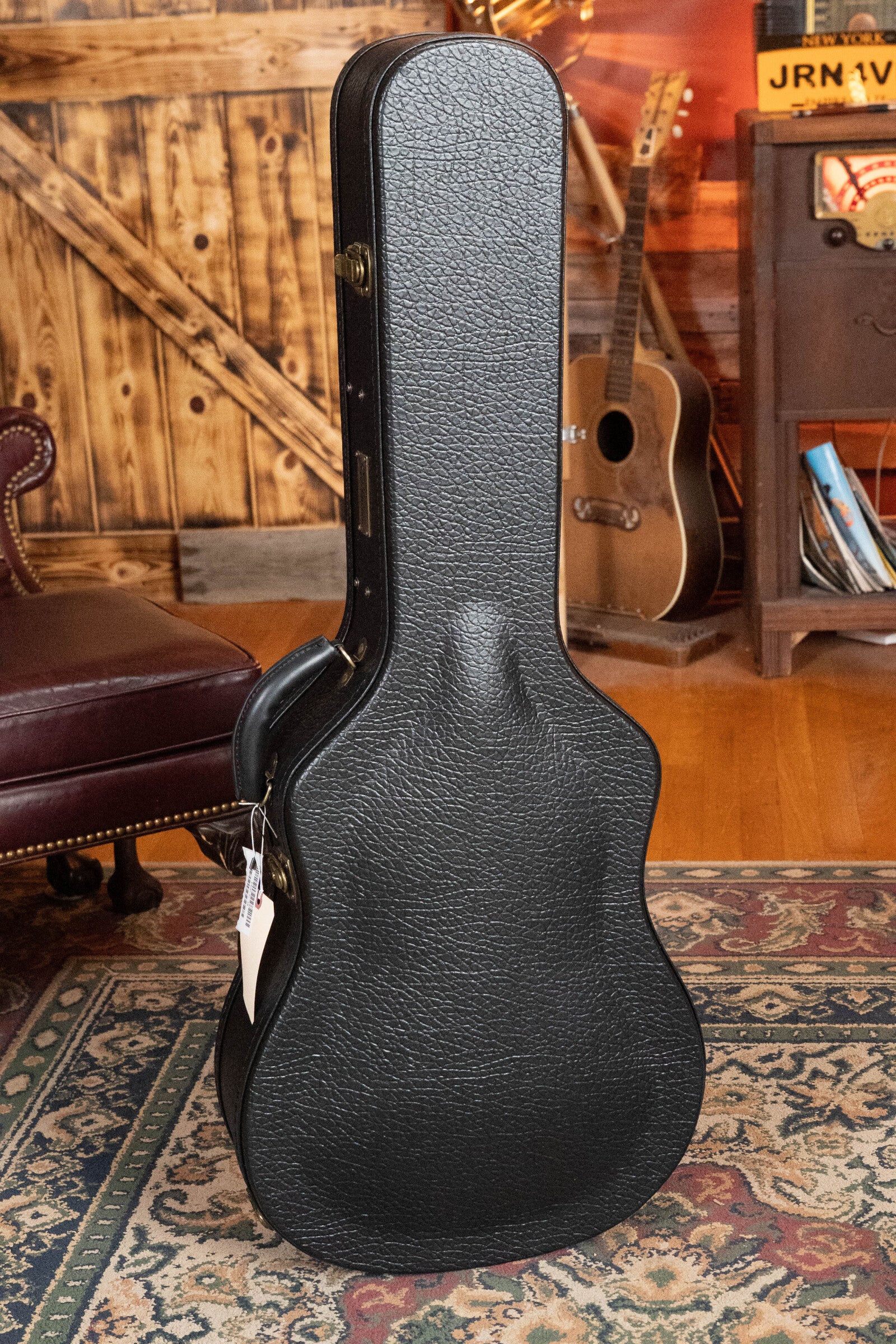 A black textured guitar case stands on a patterned rug in a cozy room, with an Eastman Guitars E40D-TC-SB Dreadnought Acoustic Guitar #1113—featuring thermo-cured AAA Adirondack top and rosewood back/sides—in the background.