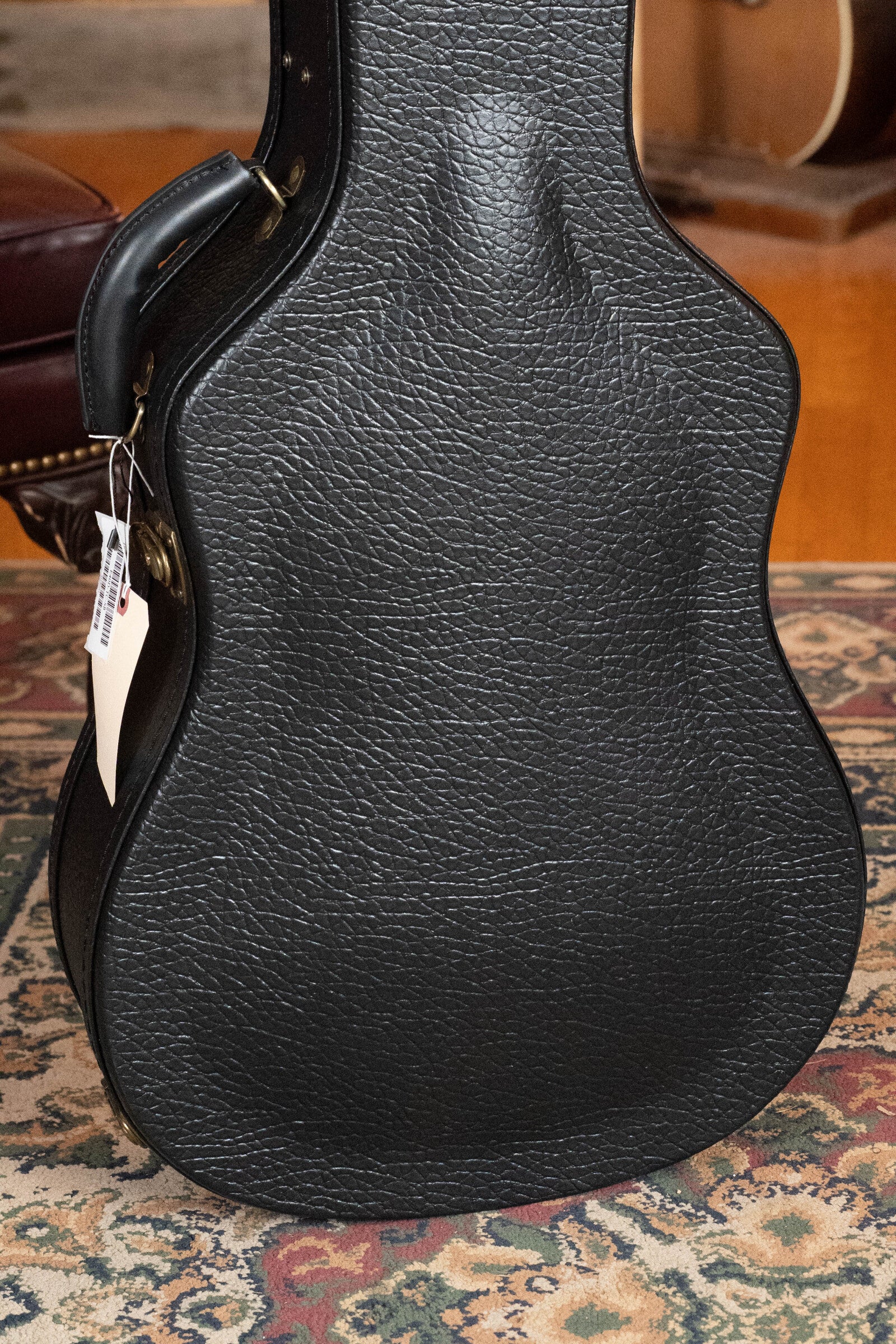 An upright black textured guitar case with handle and tag sits on a patterned rug, next to an Eastman Guitars E40D-TC-SB Sunburst Dreadnought Acoustic Guitar (#1113) featuring rosewood back and sides, with furniture in the background.