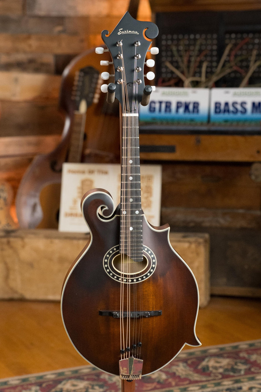 The Eastman Guitars MD314 F-Style Oval Hole Mandolin #4823, featuring a solid spruce top and ornate details, stands upright in a cozy rustic room with wooden walls, surrounded by musical instruments and sheet music.