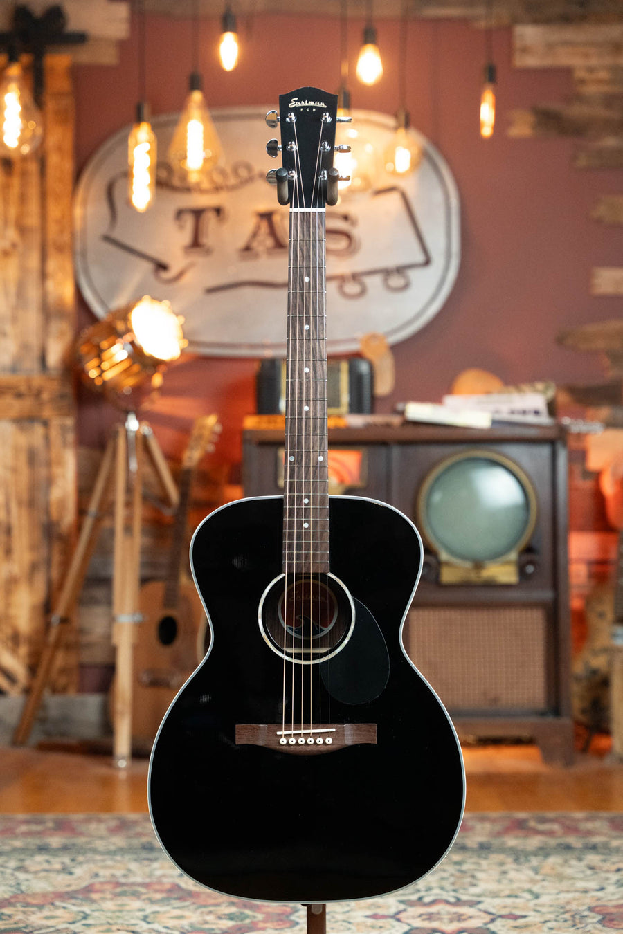 Glossy black acoustic guitar with rosewood fretboard and pearl inlays, standing in warm, rustic room with vintage decor. - Eastman PCH2-OM-BK Solid Thermo-Cured Sitka/Laminated Rosewood Orchestra Model Gloss Black Top Acoustic Guitar #1078 - Default Title