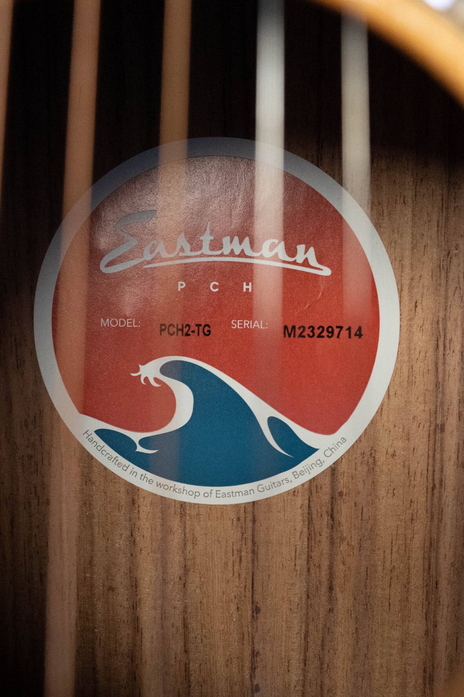 Close-up of an Eastman Guitars PCH2-TG Thermo-Cured Travel Acoustic Guitar (#9714) interior label, showing the logo, model number, serial M2329714, wave graphic, and noting it was handcrafted in Beijing, China.