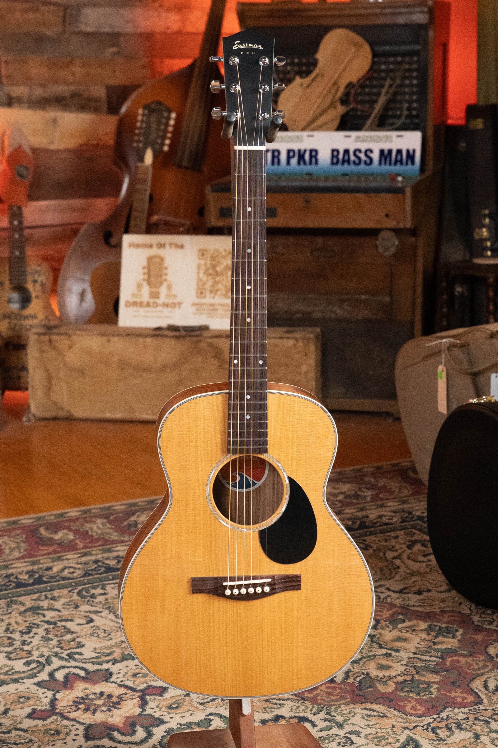 The Eastman PCH2-TG Thermo-Cured Solid Sitka/Laminated Rosewood Travel Acoustic Guitar #9714 by Eastman Guitars with a natural finish is displayed on a stand in a cozy, vintage-themed room with musical instruments and patterned rugs.