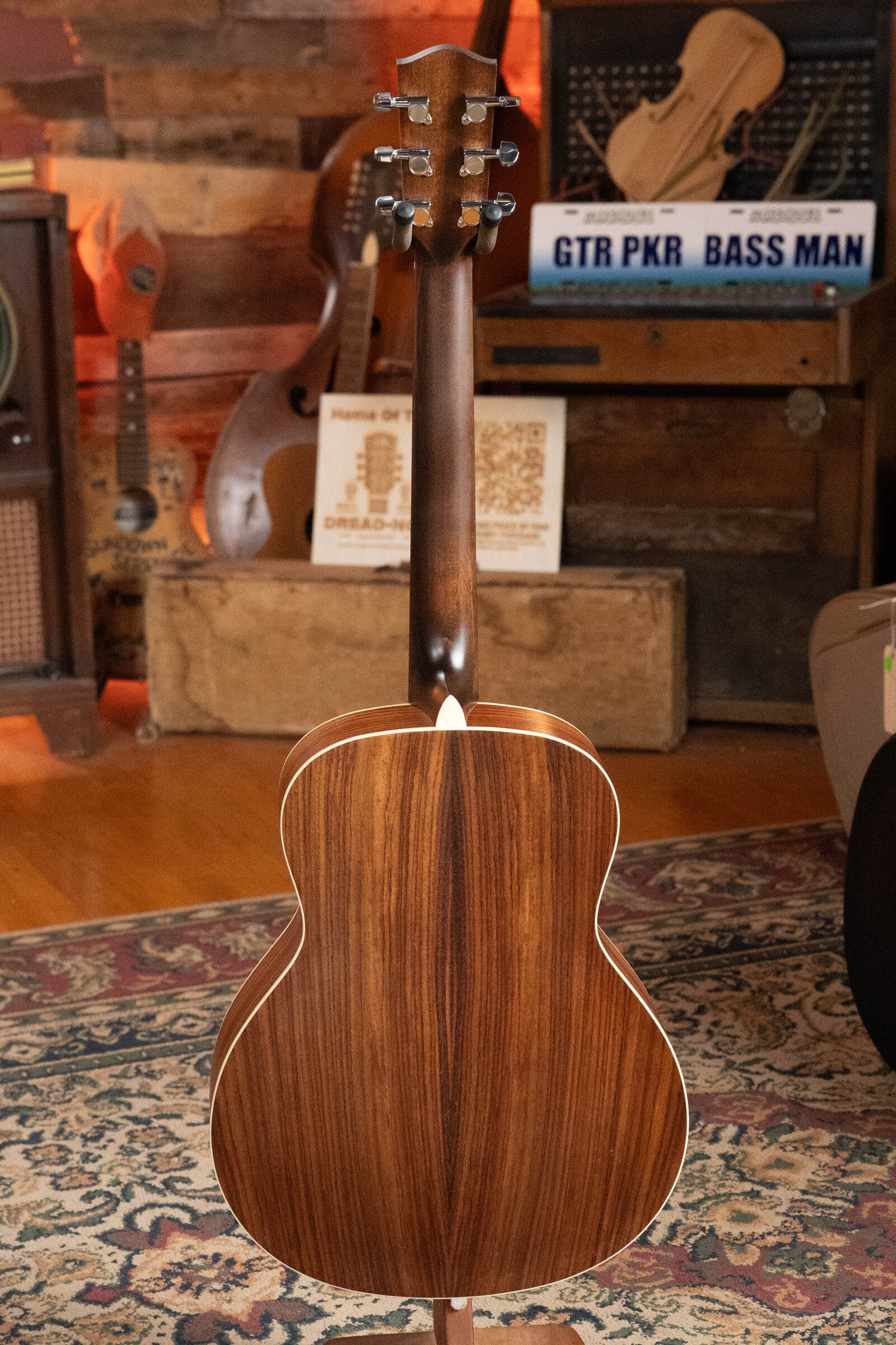 The Eastman Guitars PCH2-TG Thermo-Cured Travel Acoustic Guitar #9714 is shown from the back, highlighting its wood grain in a cozy room with other instruments, wooden walls, and music-themed decor in the background.