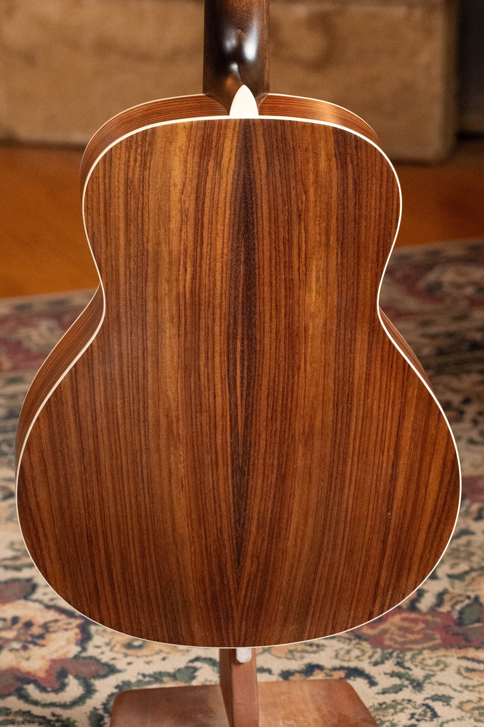 The image shows the back of the Eastman Guitars PCH2-TG Travel Acoustic Guitar (#9714) with richly grained laminated rosewood, symmetrical pattern, and white edge binding, placed on a wooden stand over a patterned rug.
