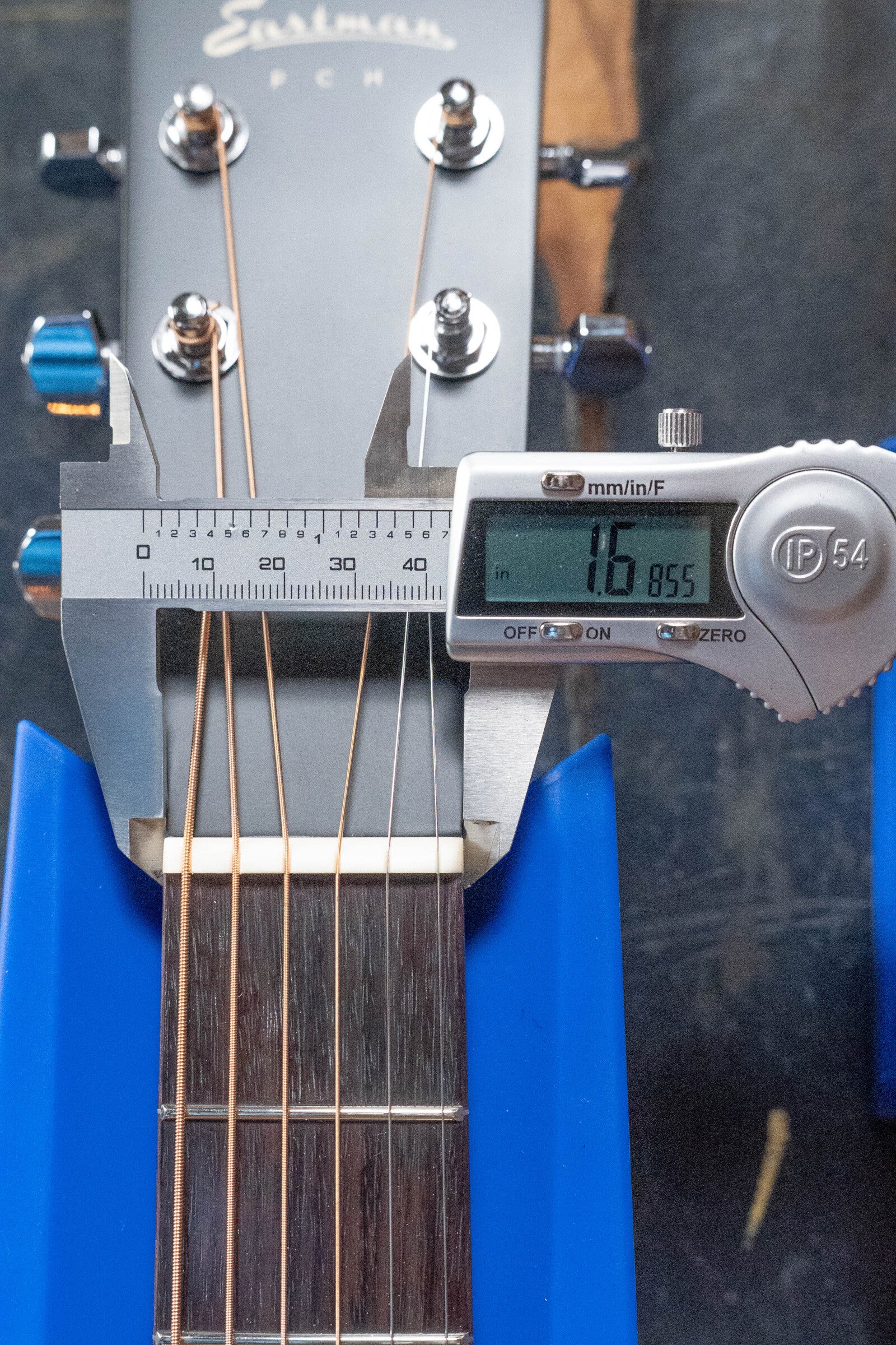 A digital caliper shows the string spacing at the nut of an Eastman Guitars PCH2-TG Travel Acoustic Guitar (#9714) as 16.85 mm, with its headstock and tuning pegs secured in a blue holder.