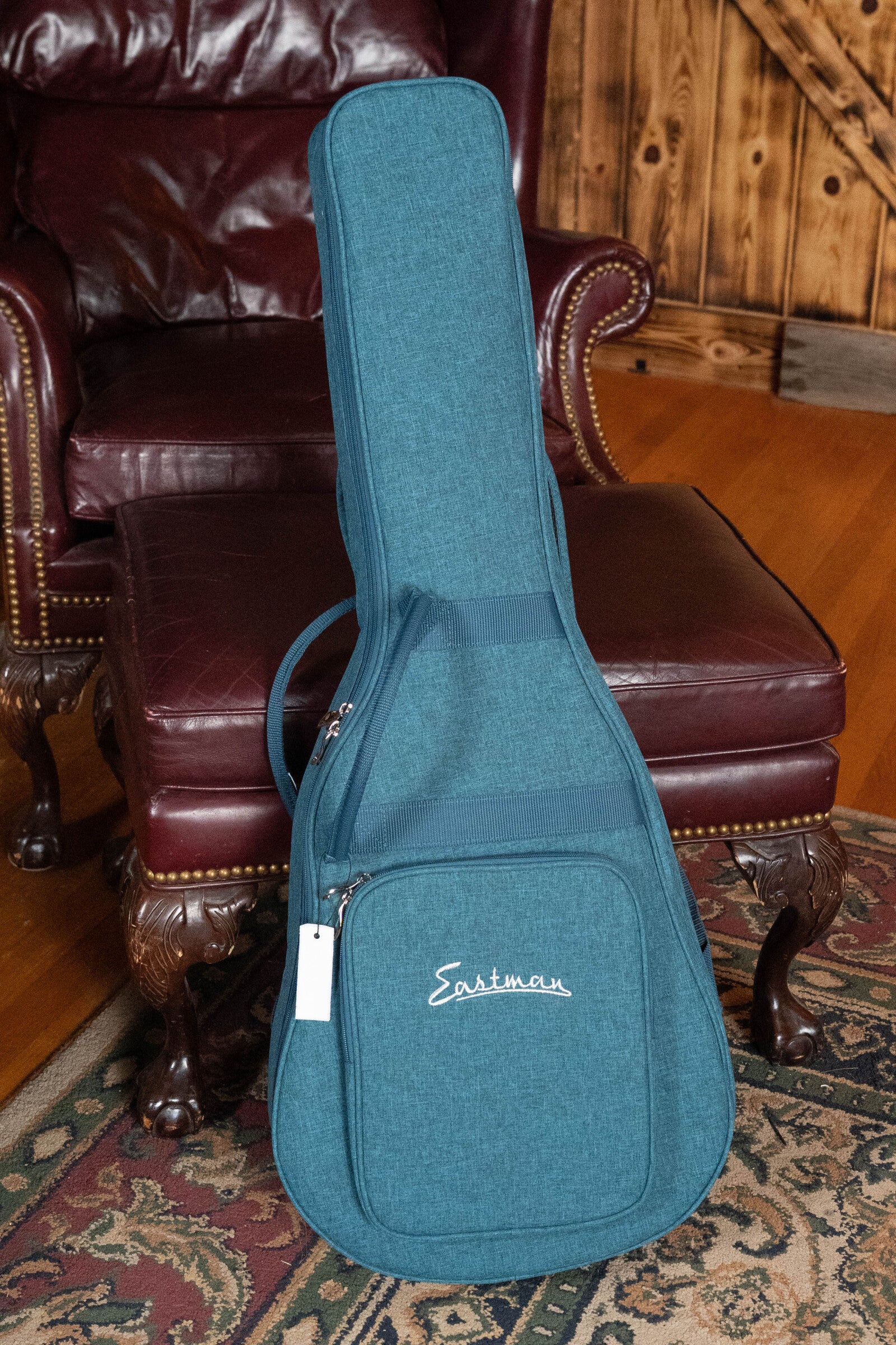 An Eastman Guitars PCH2-TG Thermo-Cured Travel Acoustic Guitar (#9714) in its teal gig bag stands upright by a maroon leather armchair with studs, on a patterned rug in a cozy wood-paneled room with hardwood floors.
