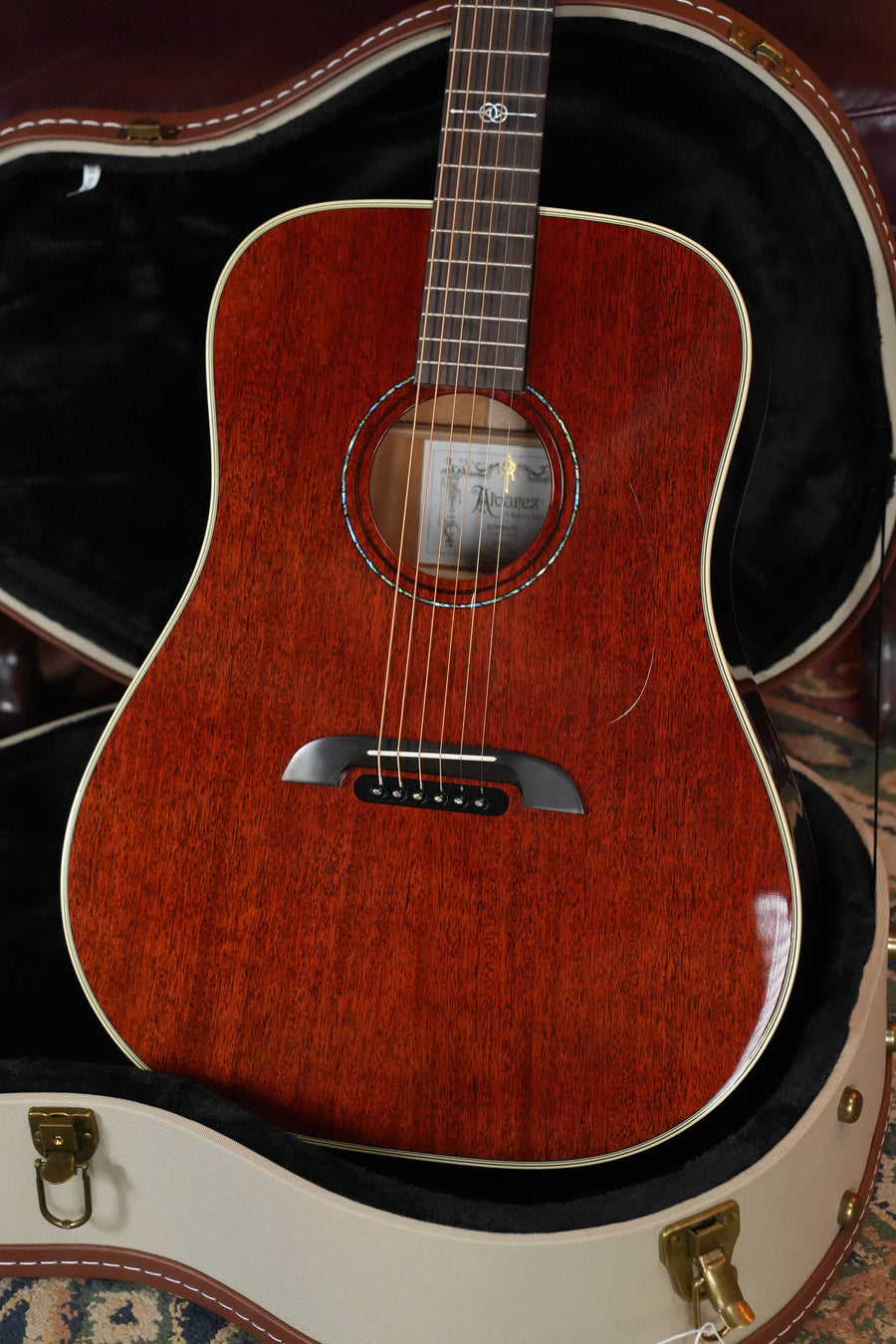 An Alvarez Yairi Honduran Series DYM66HD All Vintage Honduran Mahogany Dreadnought w/ K&K #7933 acoustic guitar in reddish-brown sits in a padded black and white hard case, showing off its glossy finish, round sound hole, and visible wood grain.