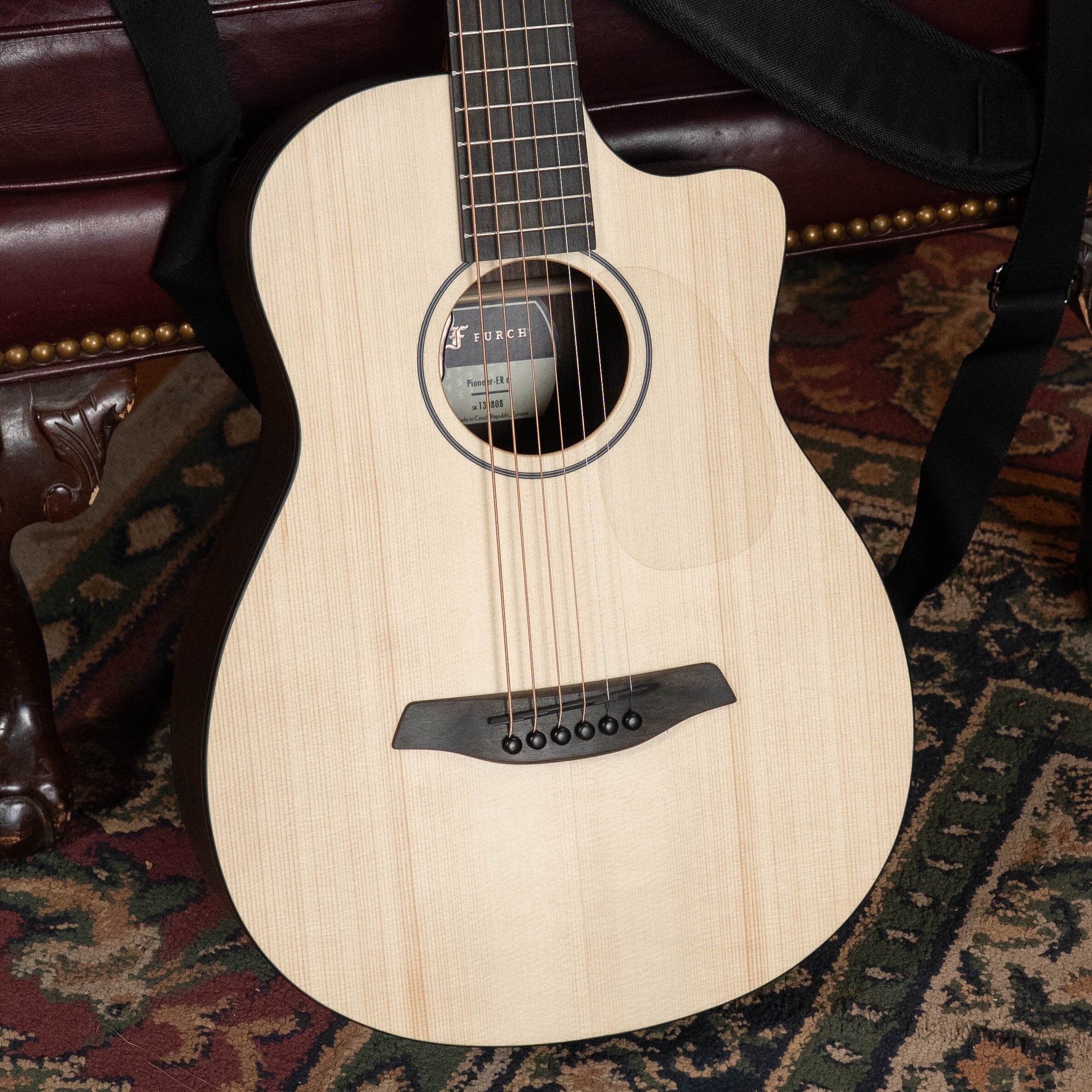 The Furch Pioneer-ER VTC Engelmann Spruce/Indian Rosewood Cutaway Travel Guitar with Soundport Boosters and VTC Pickup rests on a patterned rug and a brown leather couch with brass nailhead trim.