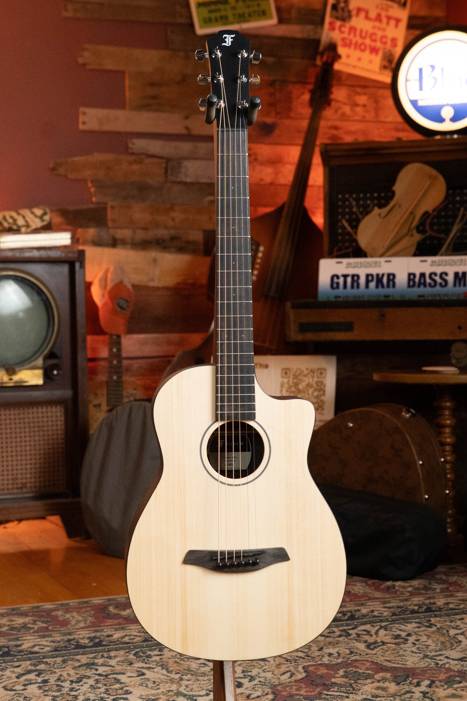 The Furch Pioneer-ER VTC Engelmann Spruce/Indian Rosewood travel guitar with Soundport Boosters and VTC Pickup stands upright in a cozy vintage room with a retro TV, wooden instruments, and music-themed signs in the background.