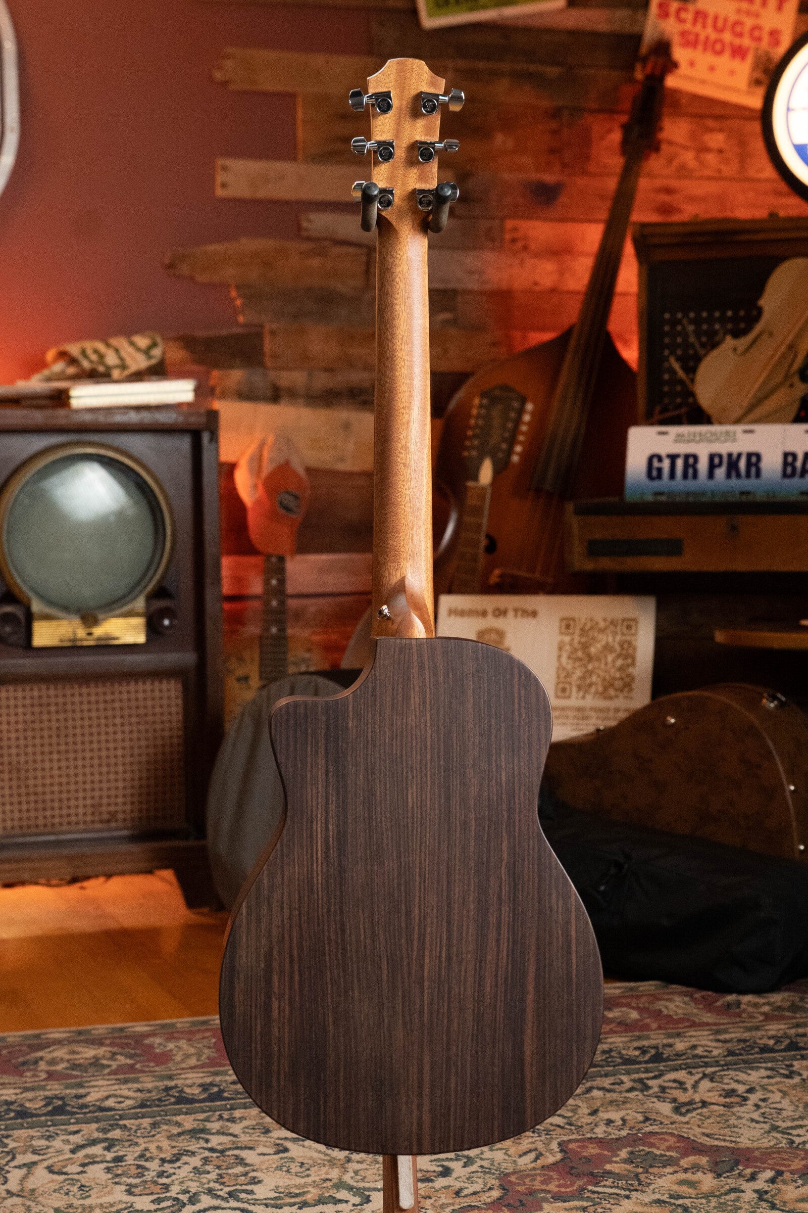 The Furch Pioneer-ER VTC Engelmann Spruce/Indian Rosewood Cutaway Travel Guitar w/Soundport Boosters & VTC Pickup #0808 stands upright in a cozy, vintage-themed room with an old TV, musical instruments, and retro signs.