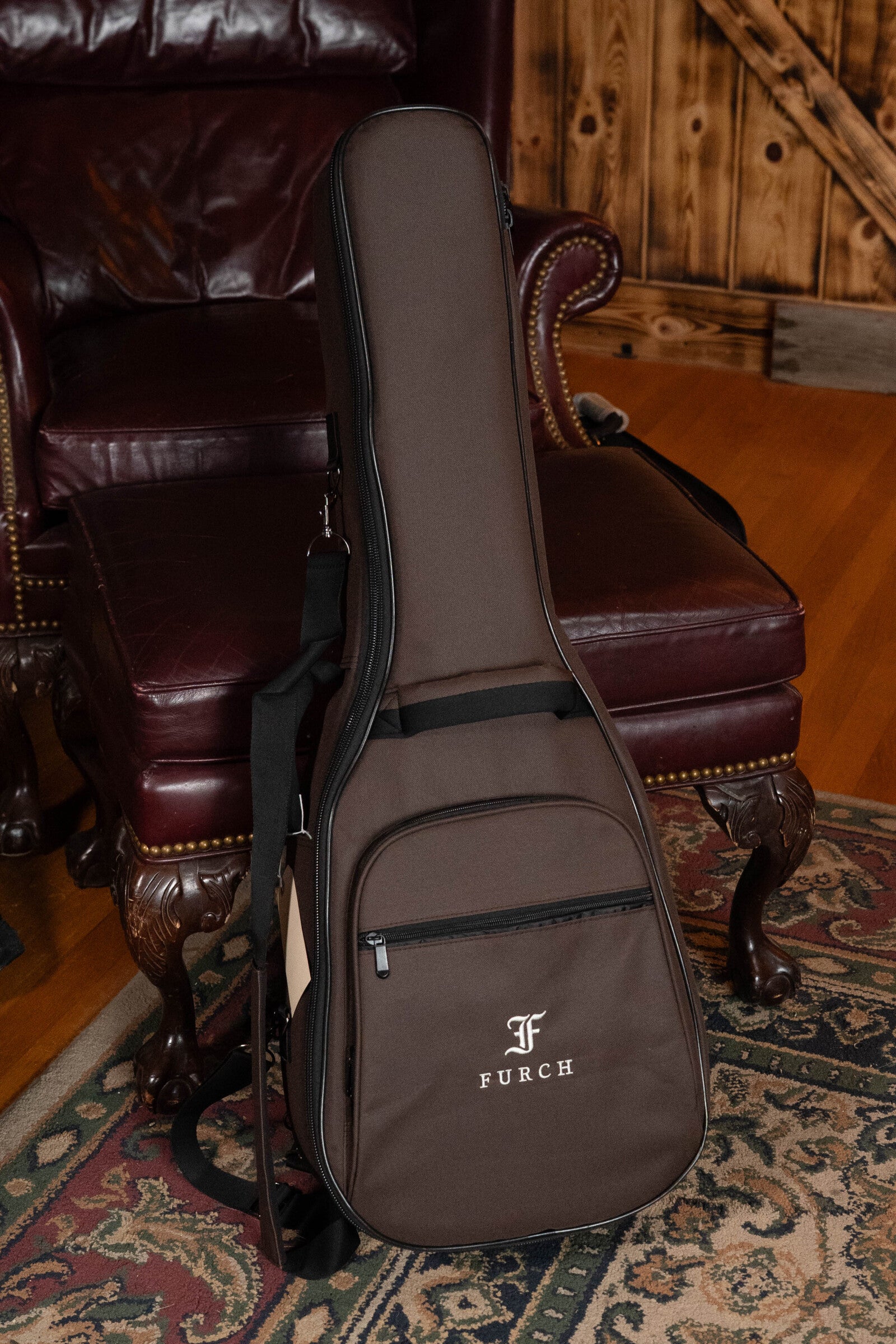 A brown Furch case for the Furch Pioneer-ER VTC Engelmann Spruce/Indian Rosewood Cutaway Travel Guitar with Soundport Boosters & VTC Pickup (#0808) stands upright on an ornate rug before a leather chair and wooden wall.