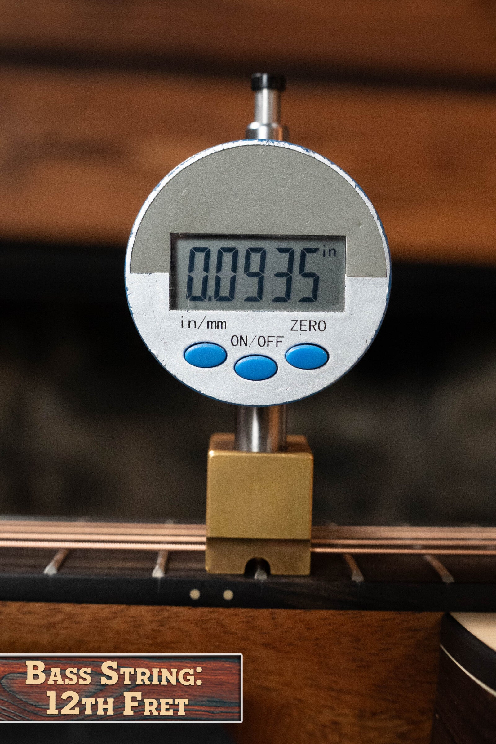 A digital gauge shows the bass string height at the 12th fret as 0.0935 inches on a Furch Pioneer-ER VTC Engelmann Spruce/Indian Rosewood Cutaway Travel Guitar, with a wooden background and a sign reading BASS STRING: 12TH FRET.