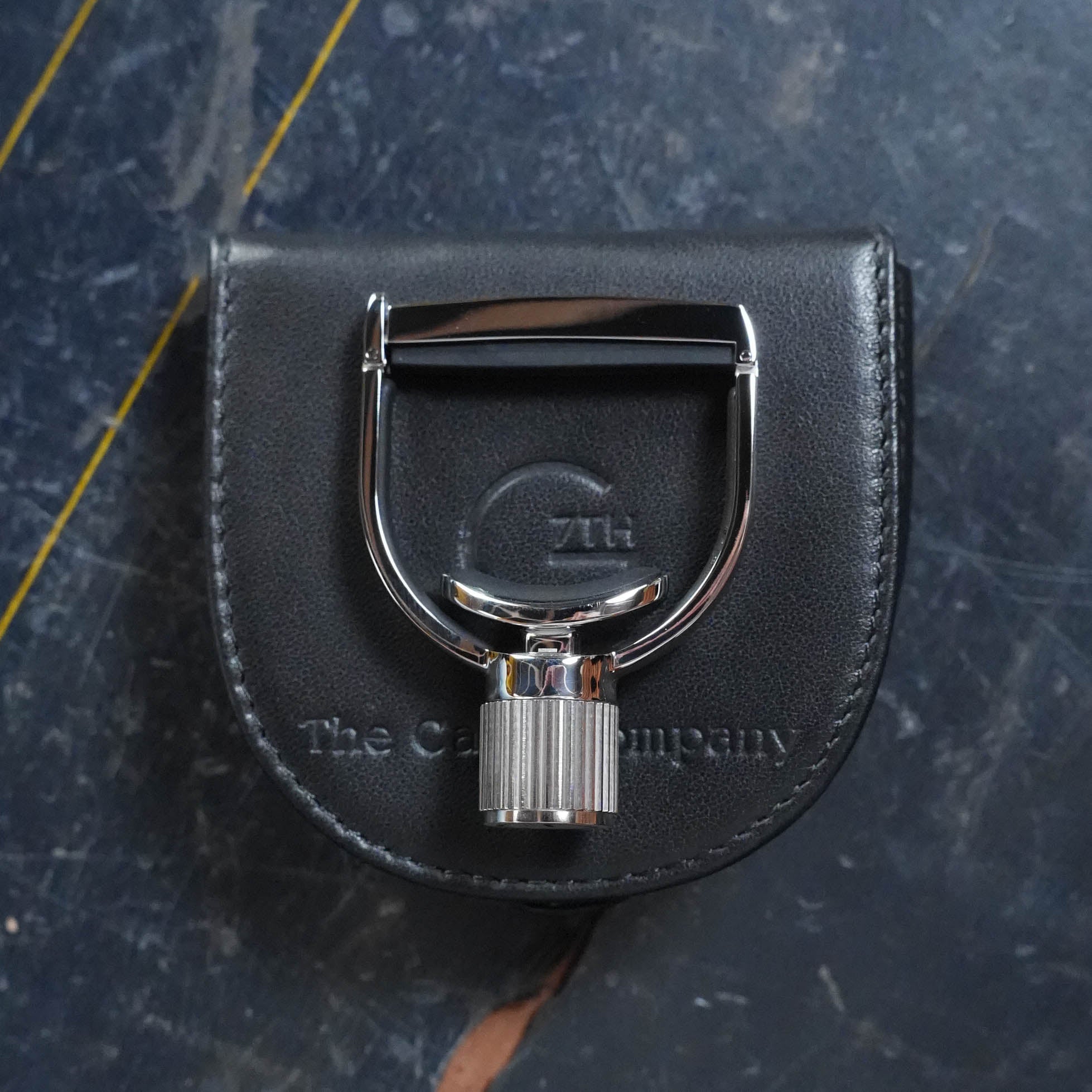 A shiny metal keychain clasp sits on a black leather pouch embossed with “The Acoustic Shoppe,” perfect for fans of the G7th Heritage Banjo Capo S1 Standard Neck Width. The set rests on a dark, textured surface with yellow diagonal lines.