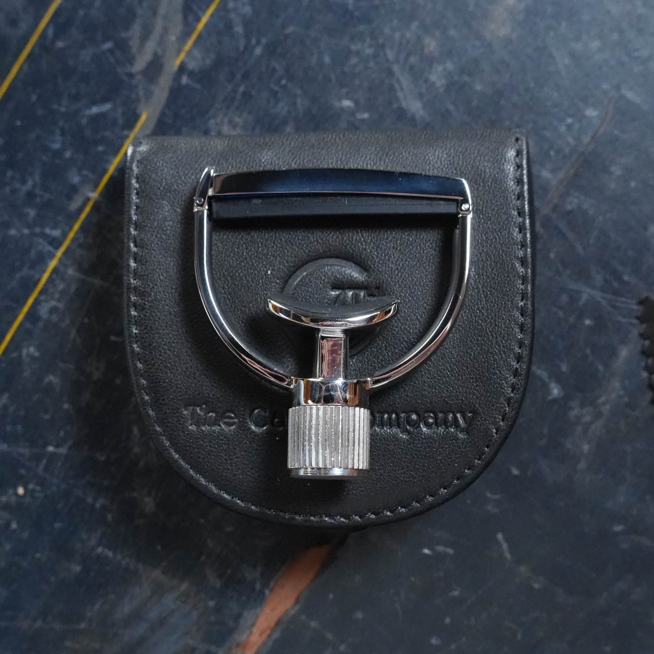 A G7th Heritage Banjo Capo S1 Wide from The Acoustic Shoppe rests on a black leather pouch embossed with the store's name, set atop a dark, textured surface with subtle yellow lines.