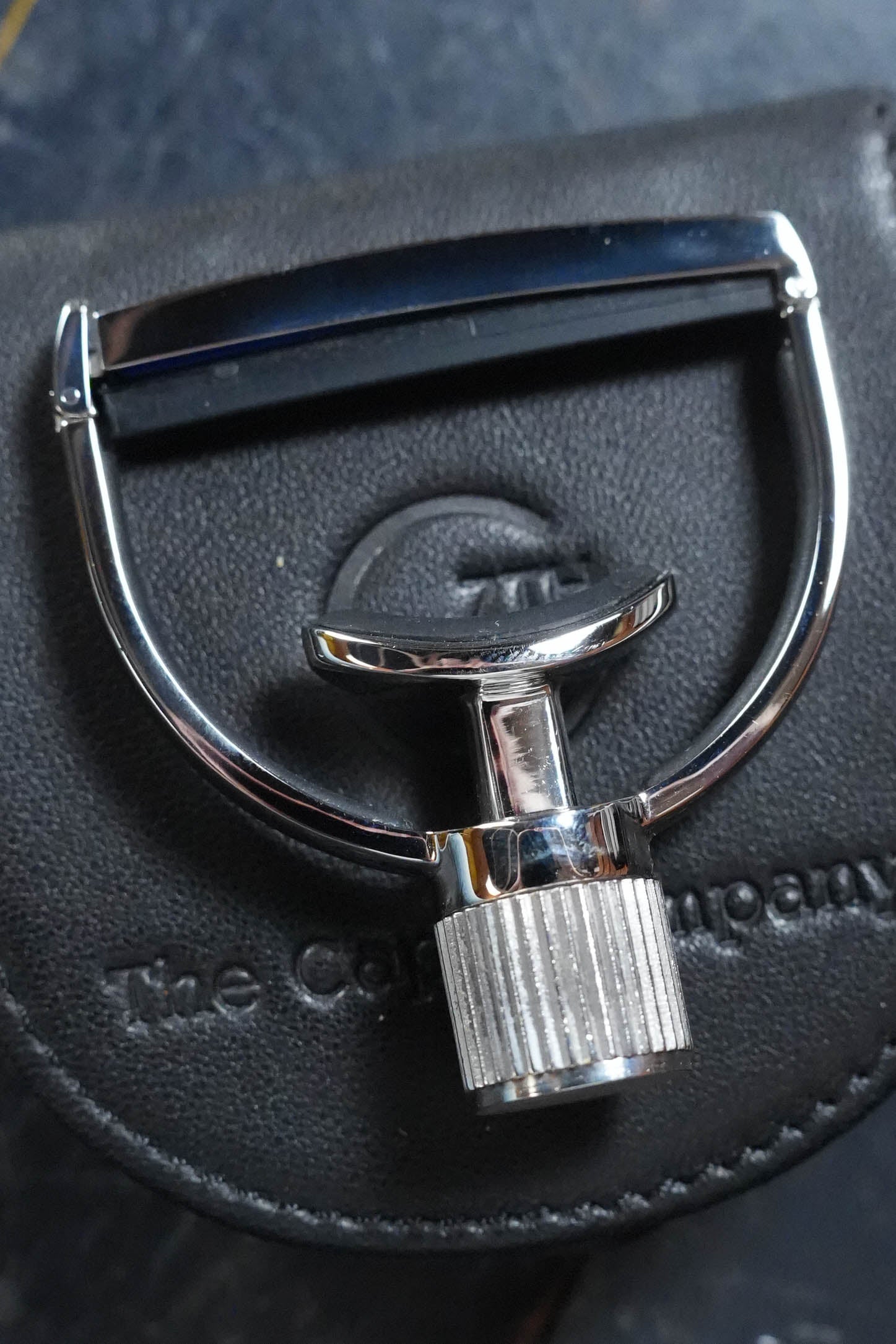 A chrome-finished G7th Heritage Banjo Capo S1 Wide by The Acoustic Shoppe rests on a black leather case embossed with “The Comp… Company.” Its yoke-style capo design features a textured handle.