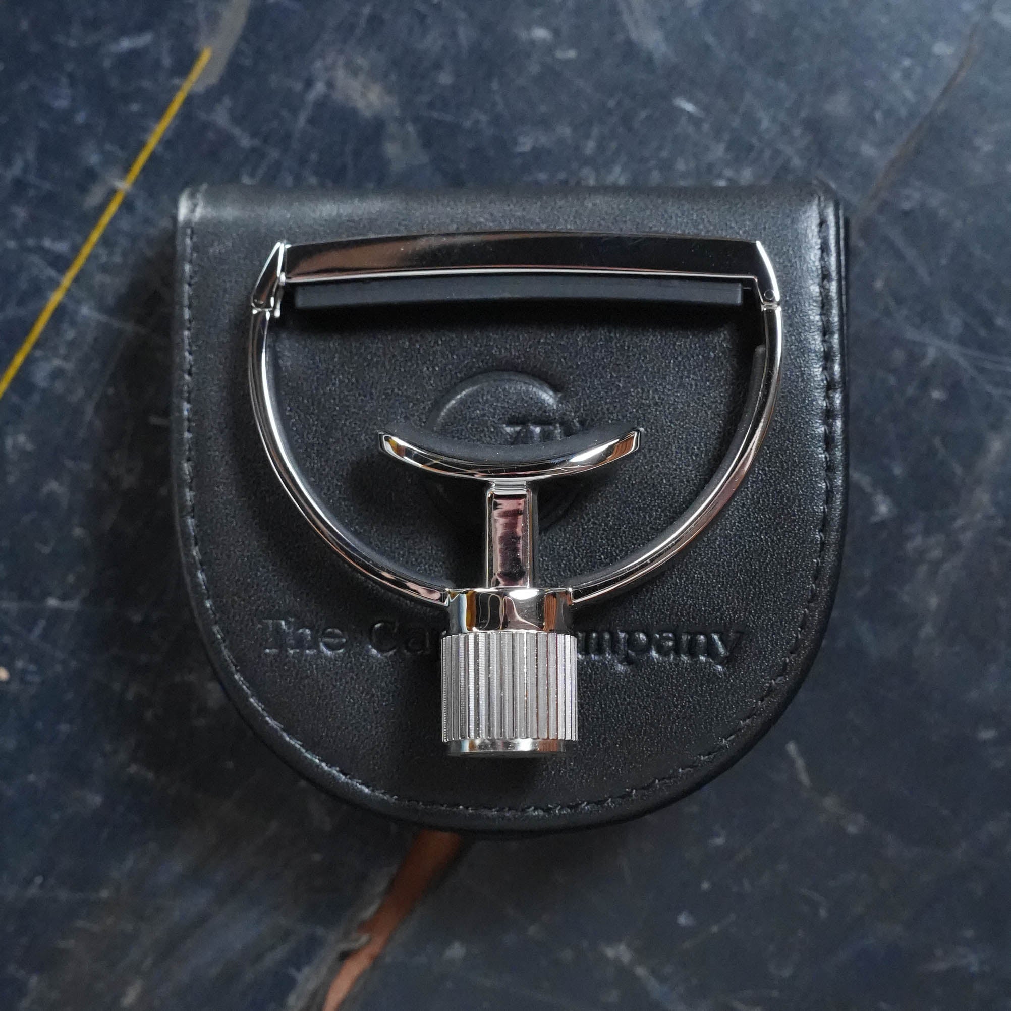 A G7th Heritage Guitar Capo S1 Standard Neck Width by The Acoustic Shoppe rests on a black leather pouch with the brand’s name, set atop a dark, textured surface marked by light scratches and a yellow line.