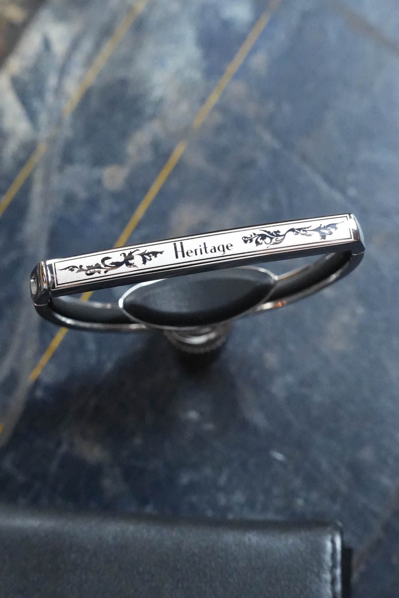A close-up of a silver bracelet engraved with "Heritage" lies beside The Acoustic Shoppe’s G7th Heritage Guitar Capo S3 Standard Neck Width, featuring Adaptive Radius Technology, both set on a dark, marble-like surface.