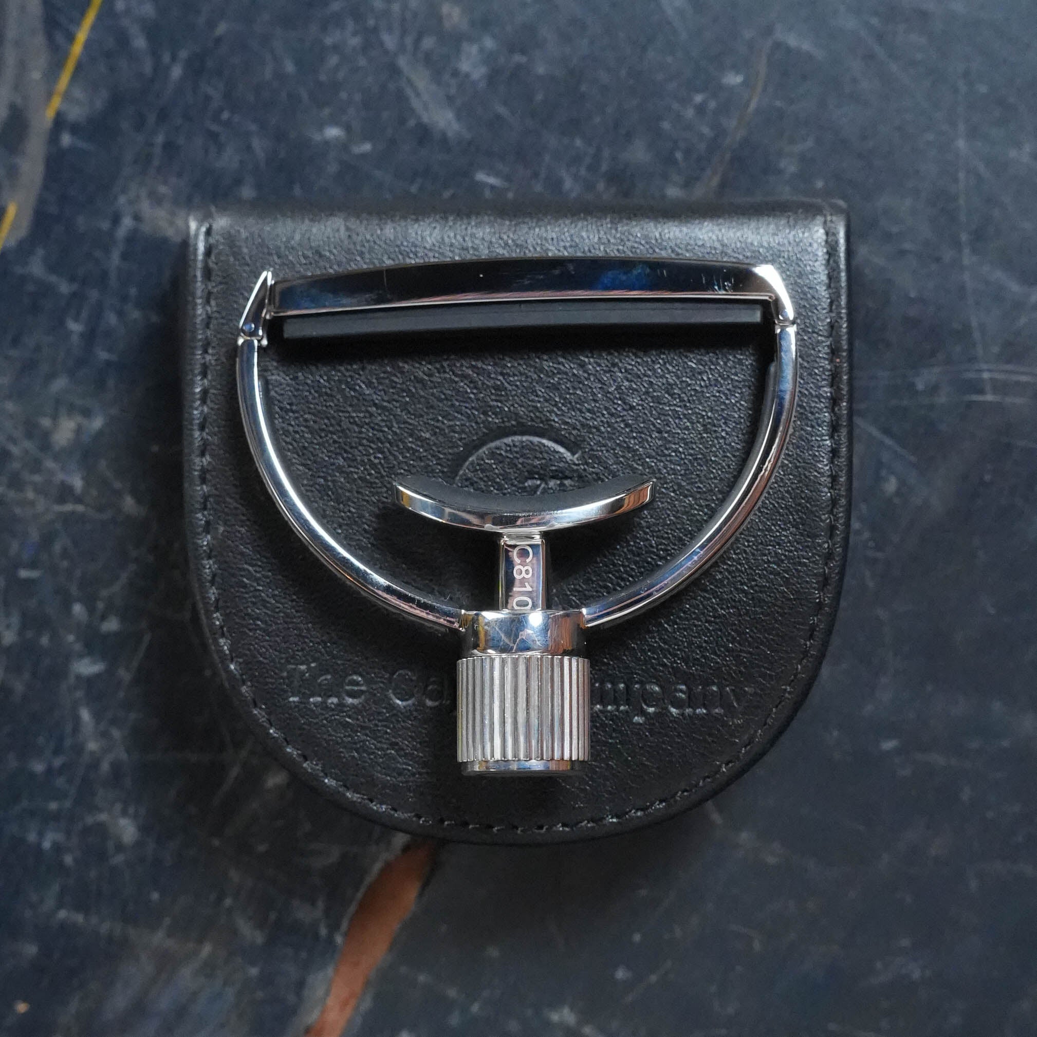 The G7th Heritage Guitar Capo S3 Wide Engraved from The Acoustic Shoppe, featuring Adaptive Radius Technology, rests atop a black leather pouch with embossed text on a dark, slightly scratched surface.