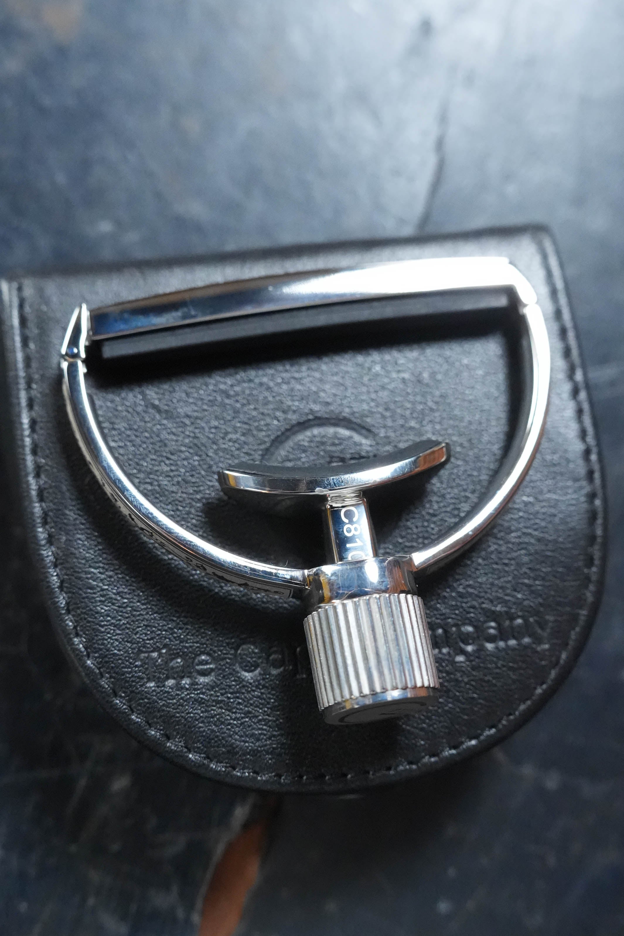 A close-up of a shiny G7th Heritage Guitar Capo S3 Wide Engraved with Adaptive Radius Technology by The Acoustic Shoppe, resting on a black leather pouch with partially visible embossed text, set against a dark, textured background.