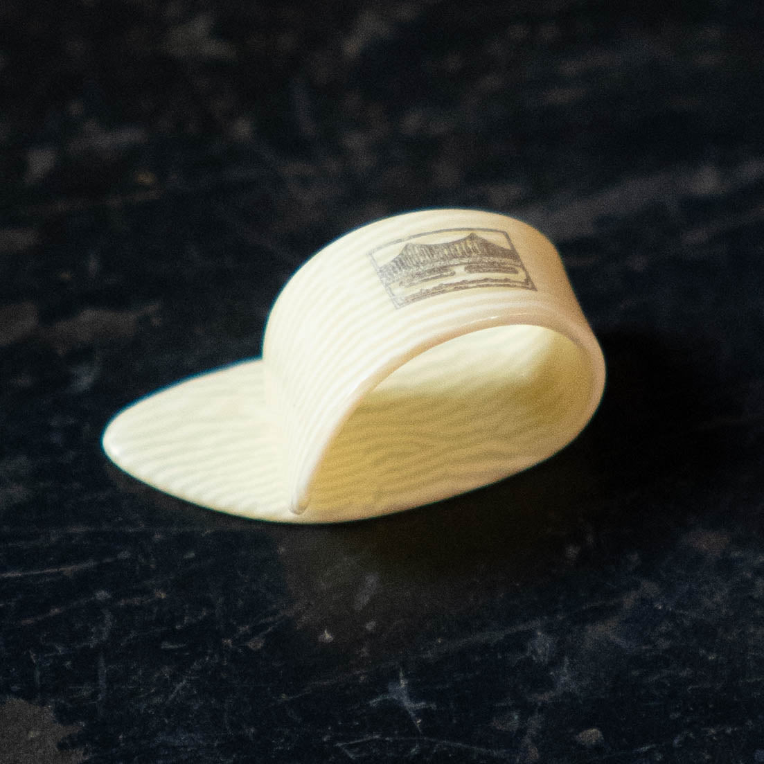 A Golden Gate GP-10 Ivoroid Thumb Pick (LARGE) by Golden Gate rests on a dark, textured surface, its faint stripes and guitar pick design adding character to its side.