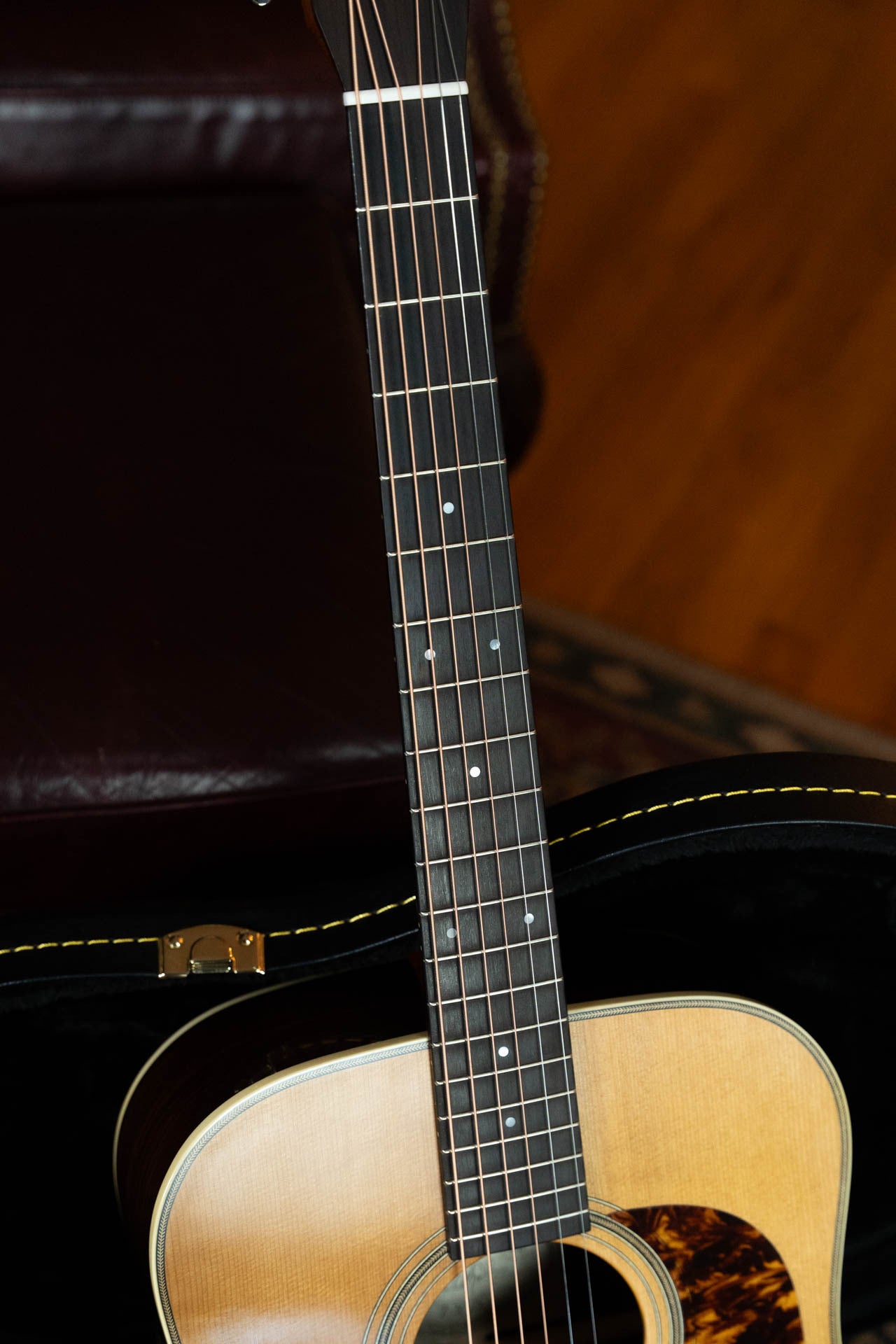 Close-up of the Gallagher Guitar Co. The Bluegrass Bell Torrefied Adirondack/Rosewood Dreadnought Acoustic Guitar #4408, showcasing its neck, fretboard, and torrefied Adirondack spruce body in a case with a brown leather chair and wood floor nearby.
