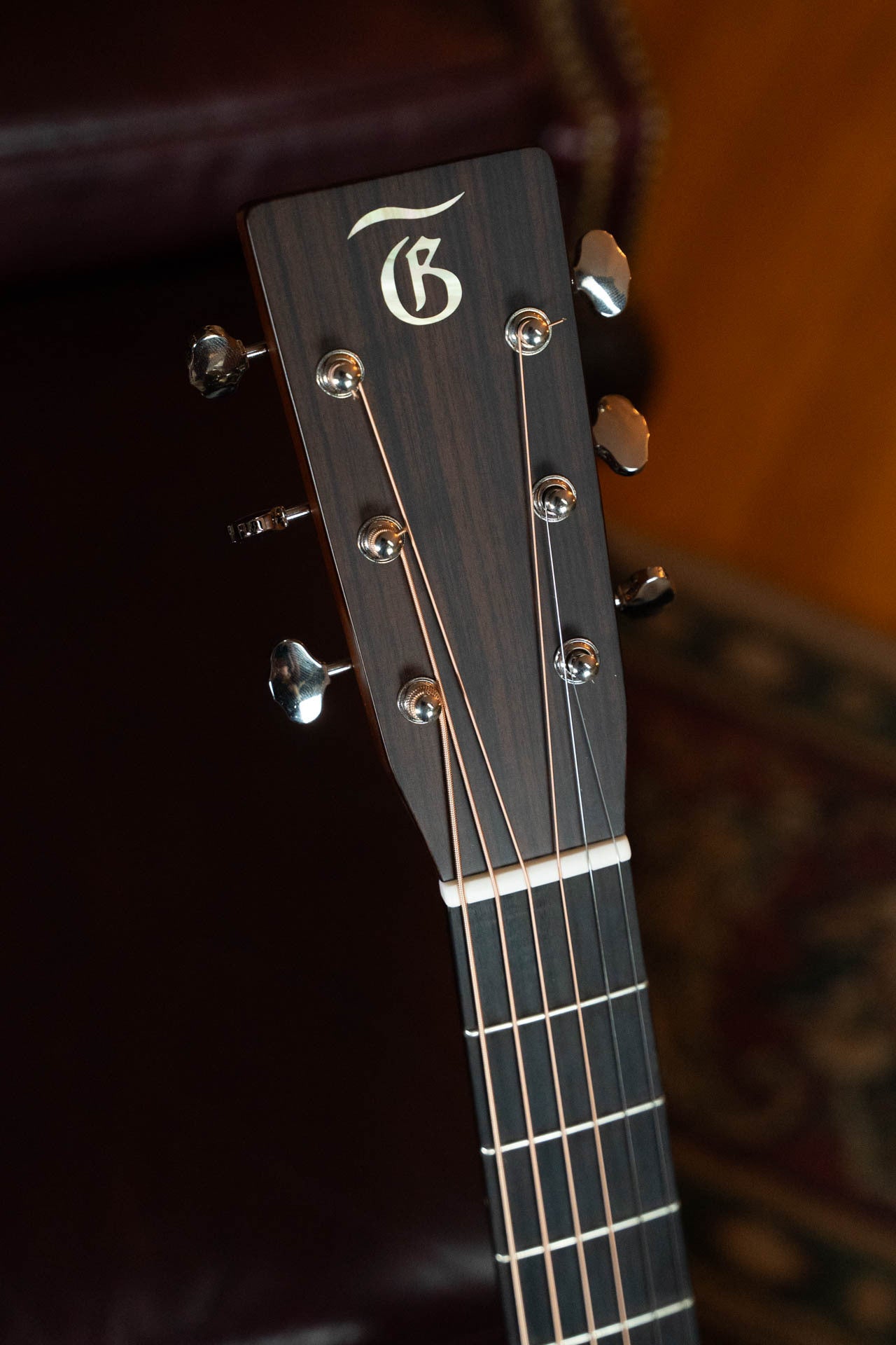 Close-up of the Gallagher The Bluegrass Bell Torrefied Adirondack/Rosewood Dreadnought Acoustic Guitar #4408 headstock by Gallagher Guitar Co., showing six tuning pegs and a stylized G logo on dark wood with metal hardware, set against a patterned surface.
