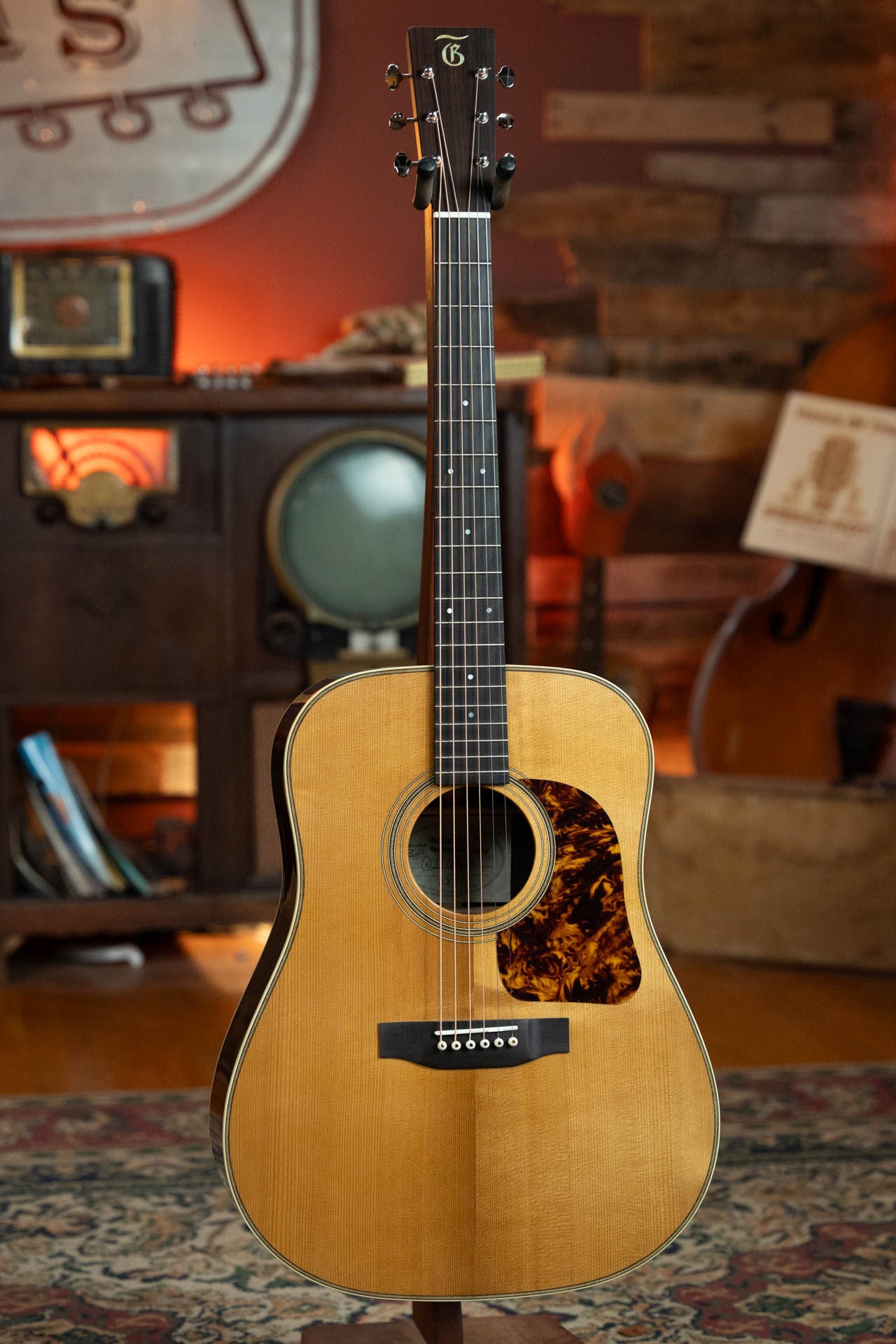 The Gallagher Guitar Co. Bluegrass Bell Torrefied Adirondack/Rosewood Dreadnought Acoustic Guitar #4408 with a natural finish and tortoiseshell pickguard stands on a patterned rug, backed by vintage radios, TV, and warm wooden decor.