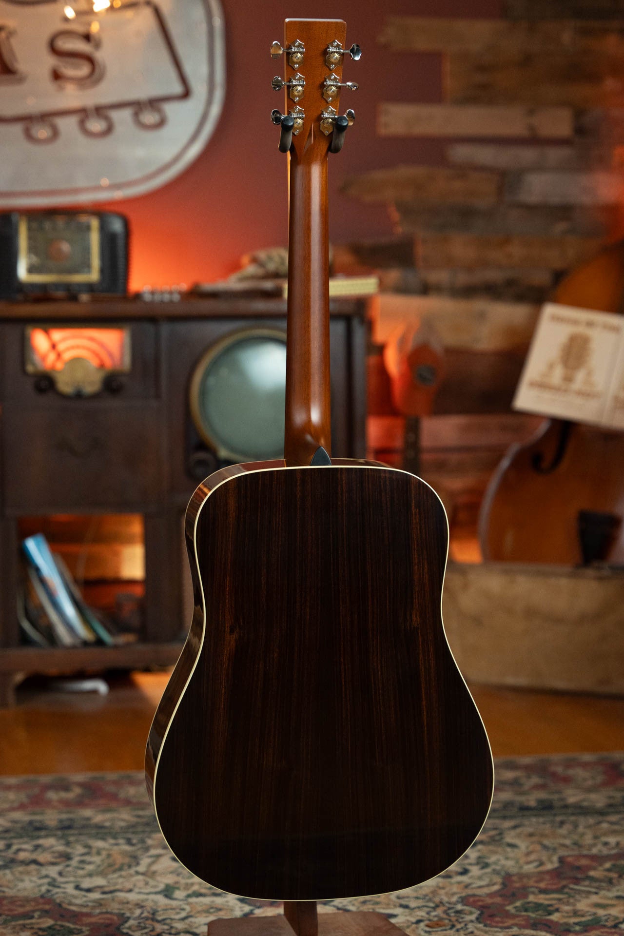 The Gallagher Guitar Co. Bluegrass Bell Torrefied Adirondack/Rosewood Dreadnought Acoustic Guitar #4408 is shown from the back, highlighting its rich wood finish amid a cozy, retro setting with a vintage radio cabinet and upright bass.