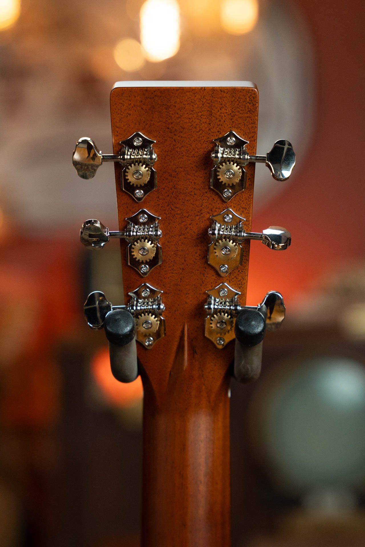 Close-up of the back of a Gallagher Guitar Co. The Bluegrass Bell Torrefied Adirondack/Rosewood Dreadnought Acoustic Guitar #4408 headstock with six metal tuners, upright on a stand against a warmly lit, blurred background.