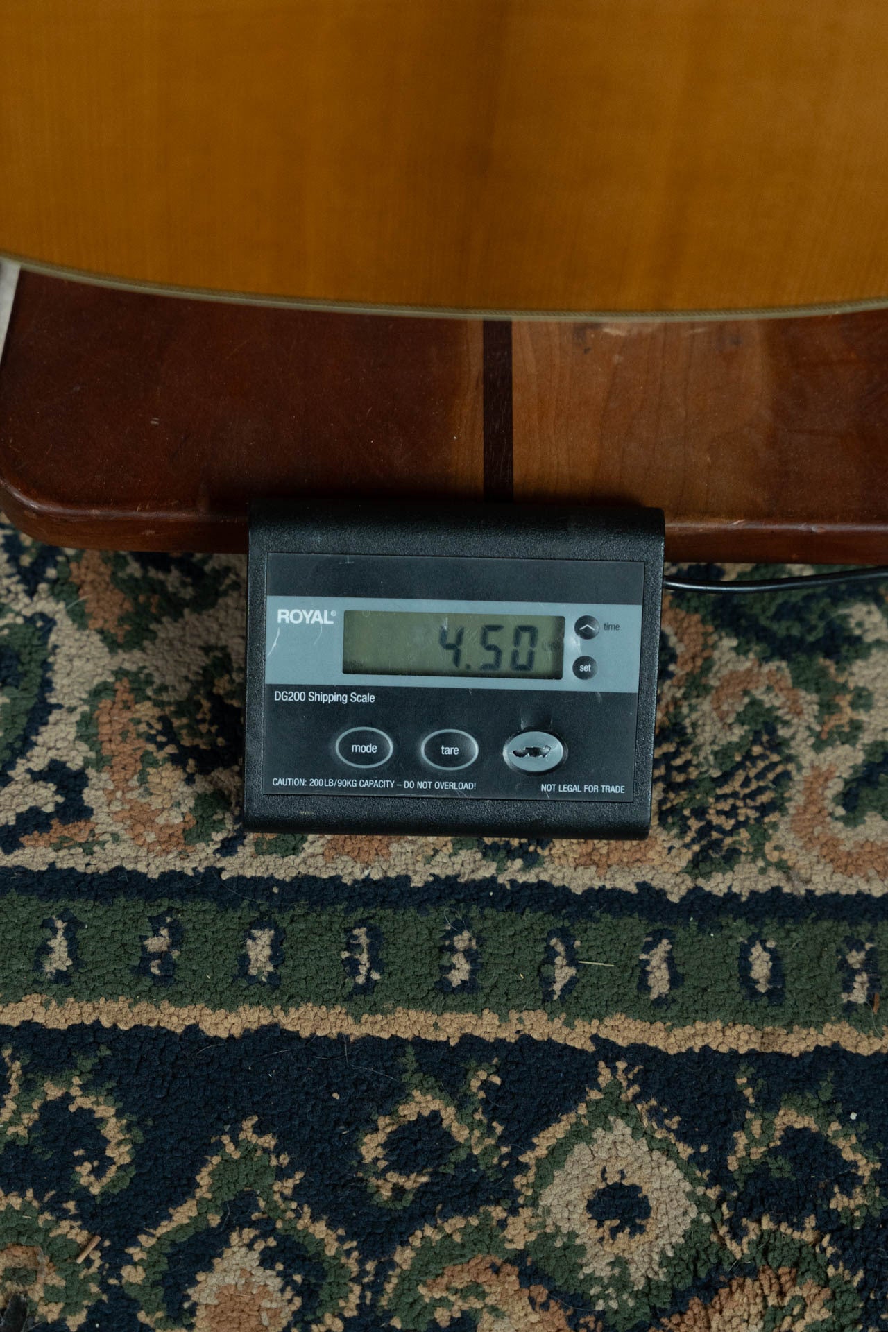 A digital shipping scale reads 4.50 pounds on a patterned carpet, with a wooden surface and the body of a Gallagher The Bluegrass Bell Torrefied Adirondack/Rosewood Dreadnought Acoustic Guitar (#4408) by Gallagher Guitar Co. in the background.