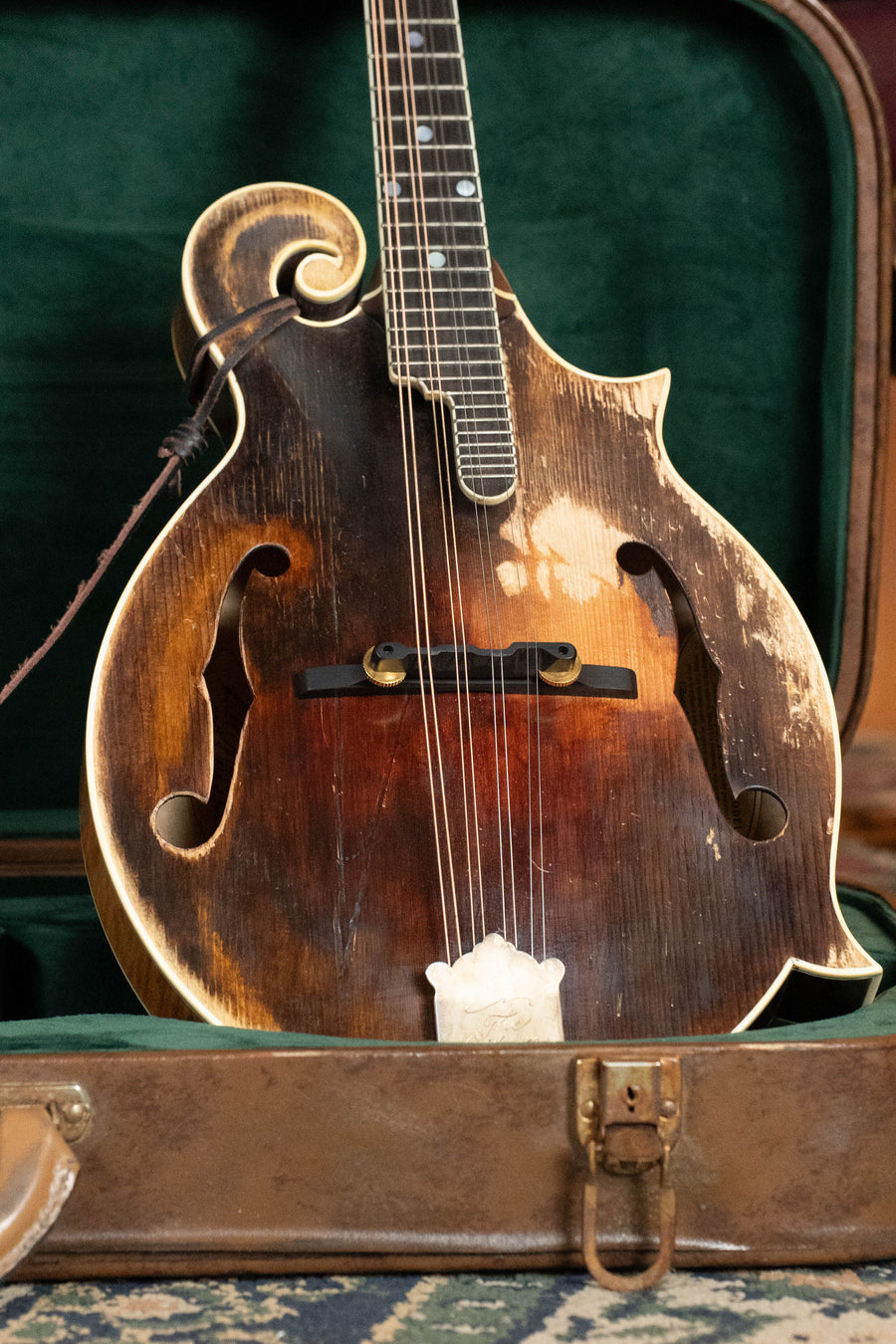 A Gibson F-5 Hall of Fame Monroe Cremona Burst Mandolin GH with a worn, vintage look and visible scratches rests in an open green-lined case, its dark sunburst finish showing areas of fading on the wooden body.