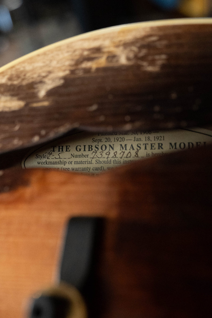 A close-up of a worn Gibson mandolin reveals a partially visible label inside reading THE GIBSON MASTER MODEL, with faded serial numbers and warranty info. The instrument is the Gibson F-5 Hall of Fame Monroe Cremona Burst Mandolin GH.