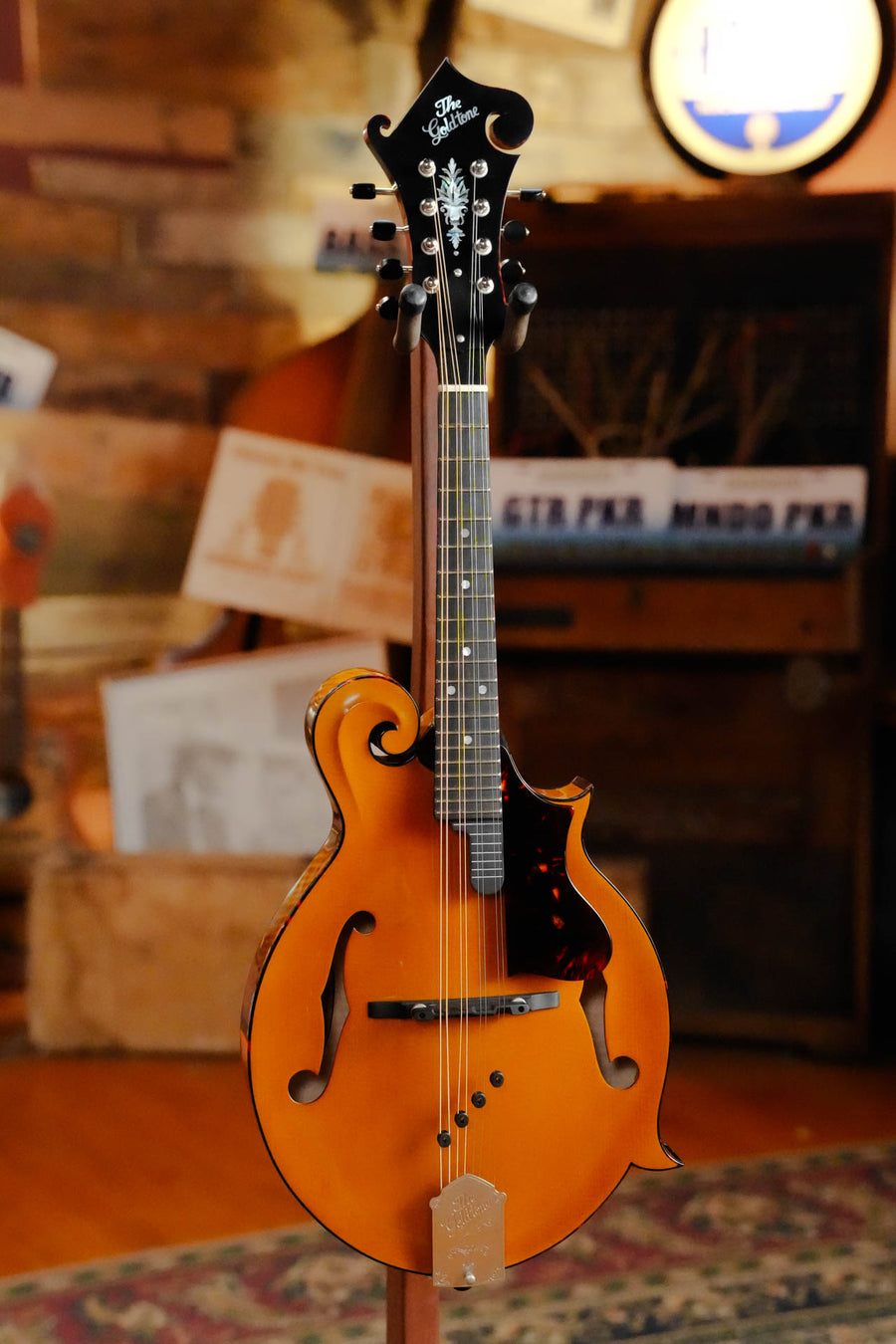 A Gold Tone Grandmaster Mandolin Elite - Honey Amber Finish #5627 stands on display in a warmly lit, rustic room; its F-style body, ornate scrollwork, premium tonewoods, and black pickguard highlight the craftsmanship of this Gold Tone instrument.