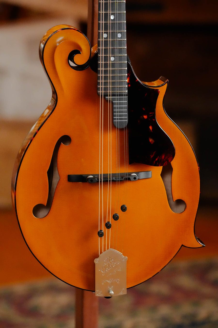 A close-up of the Gold Tone Grandmaster Mandolin Elite - Honey Amber Finish #5627, an F-style mandolin featuring a glossy honey amber finish, black pickguard, and decorative scroll by Gold Tone, standing upright indoors.