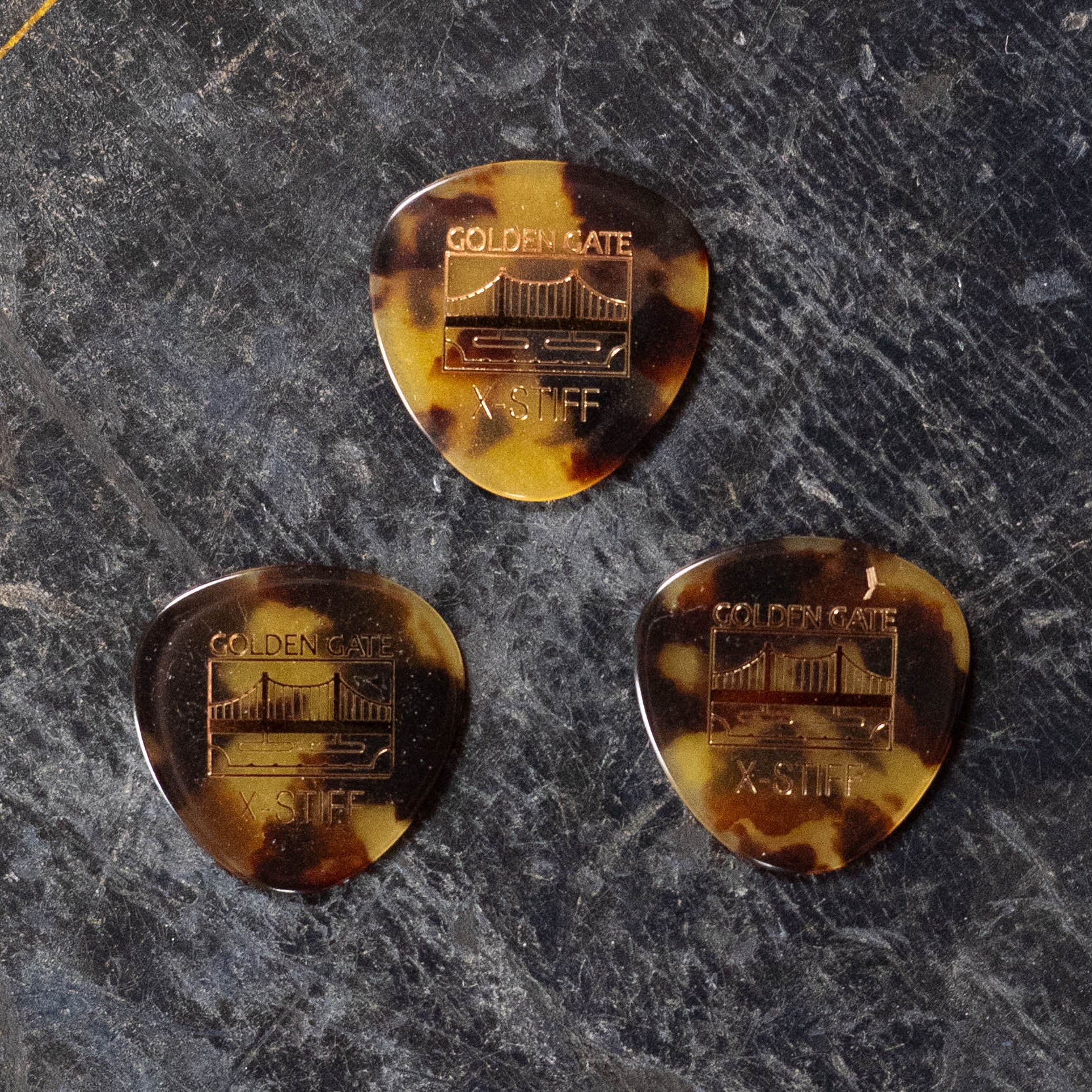 Three Golden Gate Flat Pick Round Tortoise Triangle picks (3 Pack) by Golden Gate, featuring mottled brown design and X-Stiff gauge, rest on a dark surface—perfect flat picks for rich, resonant tones.
