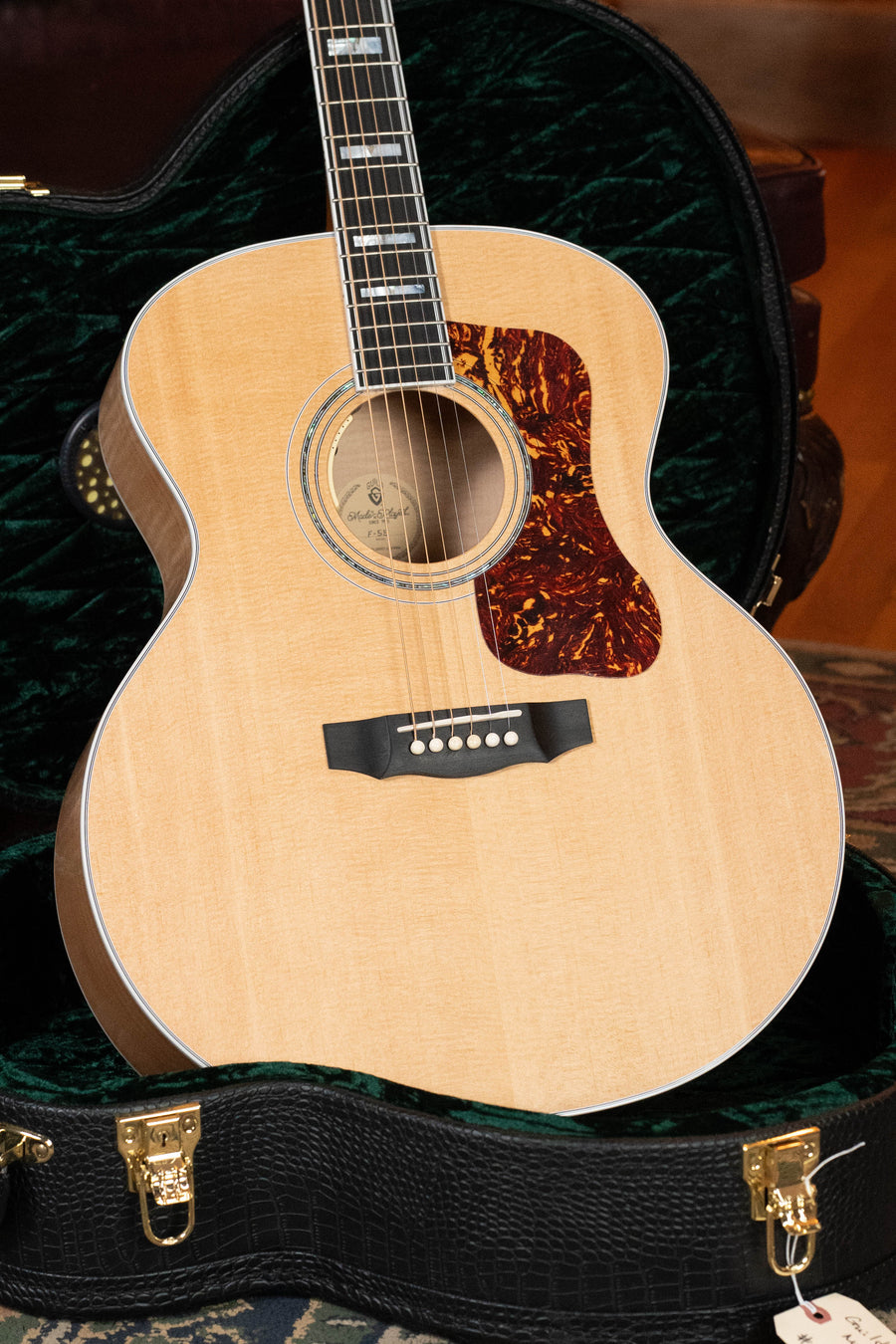 A Guild Guitars F-55E Sitka/Maple Jumbo Natural Gloss acoustic guitar with electronics (#0842) and a tortoiseshell pickguard sits upright in an open, black, velvet-lined case, showing its metal strings and sound hole.