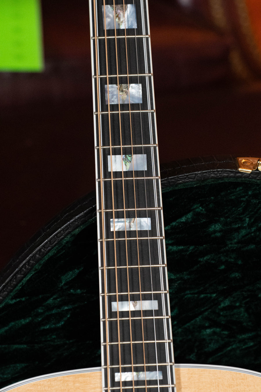 Close-up of a Guild Guitars F-55E Sitka/Maple Jumbo Natural Gloss Guitar neck with frets and mother-of-pearl inlays, plus part of the body and its green velvet-lined case visible in the background.
