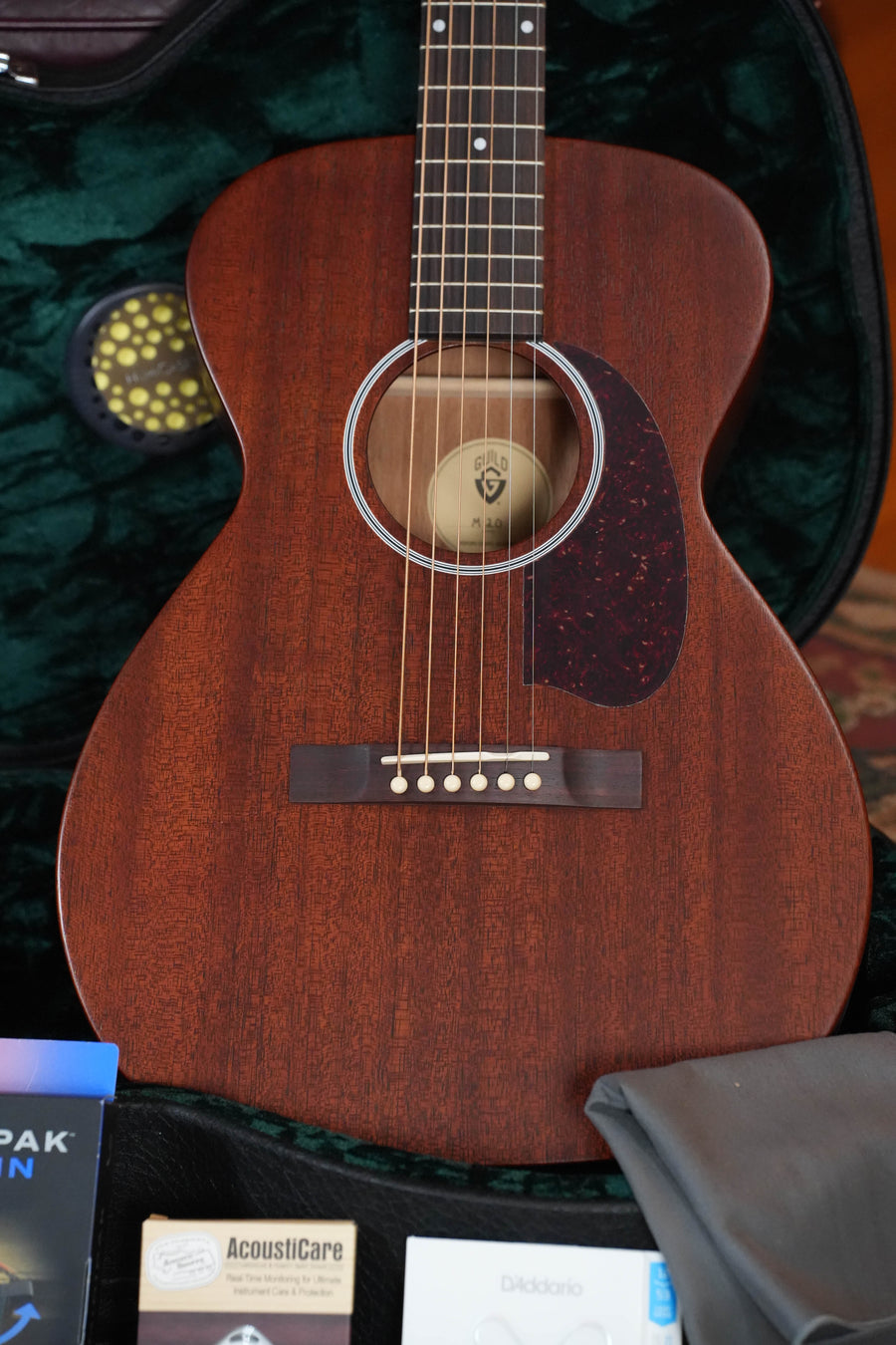 A Guild Guitars M-20 Natural All African Mahogany Concert Acoustic Guitar (#0317) with a dark pickguard rests inside a green velvet-lined case, with AcoustiCare and D’Addario accessories partially visible in front.