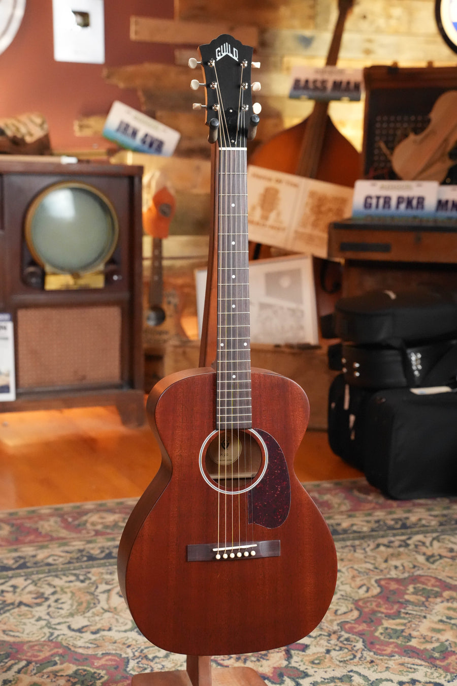 A Guild Guitars M-20 Natural All African Mahogany Concert Acoustic Guitar (#0317) rests on a stand in a cozy, vintage-themed room with an old TV, suitcases, instruments, posters, a patterned rug, and wooden floor.