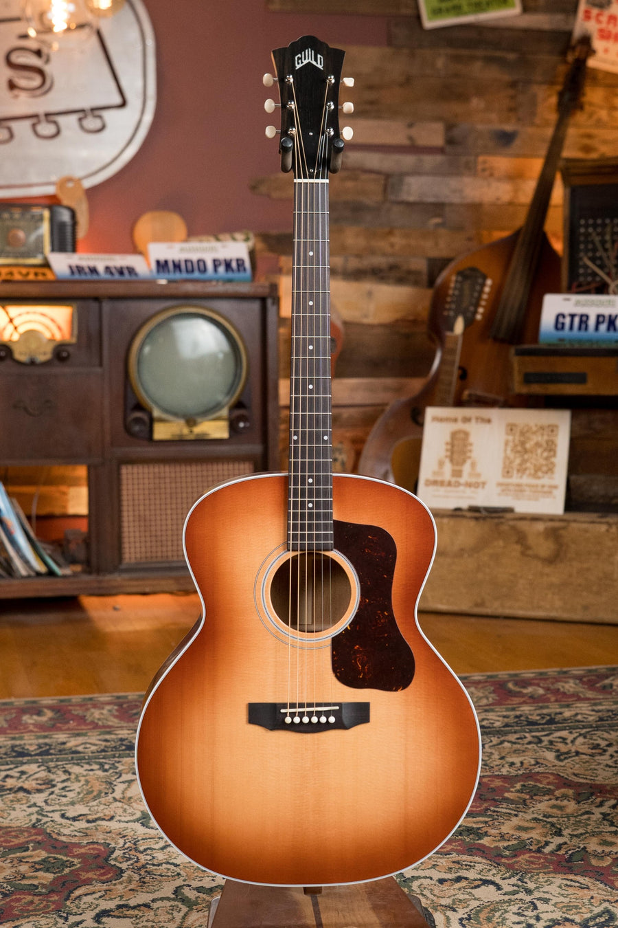 A Guild Guitars F-40 Standard Sitka/Mahogany Pacific Sunset Burst Jumbo Acoustic Guitar #0068 stands upright on an ornate rug in a vintage room with wooden walls, an old TV, and assorted musical instruments and signs.