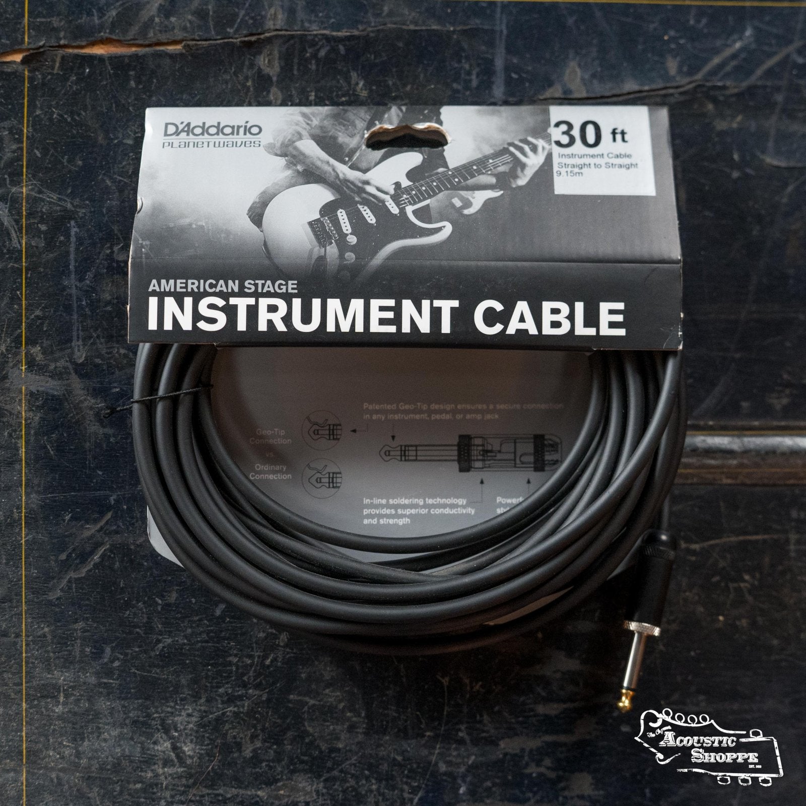 A 30-foot D'Addario PW-AMSG American Stage Instrument Cable (1/4" to 1/4") is shown coiled in its packaging, featuring a guitarist image and product details, with the “Acoustic Shoppe” logo on a dark surface.