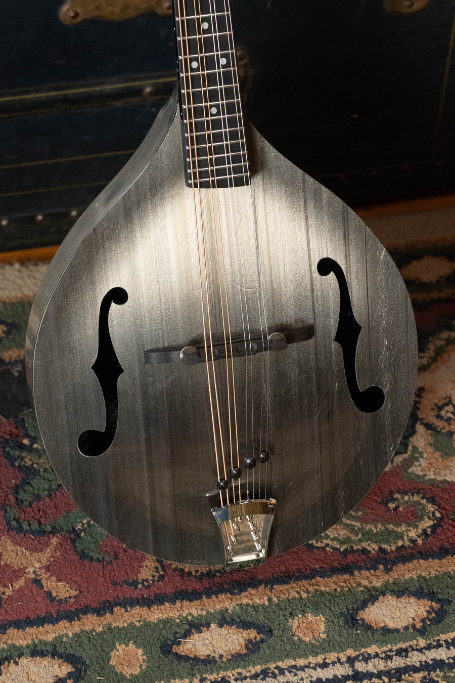 A close-up of the KLOS Guitars CT_MA_DA Full Carbon Timber A-Style Acoustic Mandolin #2680 with a metallic silver finish and f-shaped sound holes, featuring an adjustable ebony bridge, on a patterned rug near a dark wooden chest.