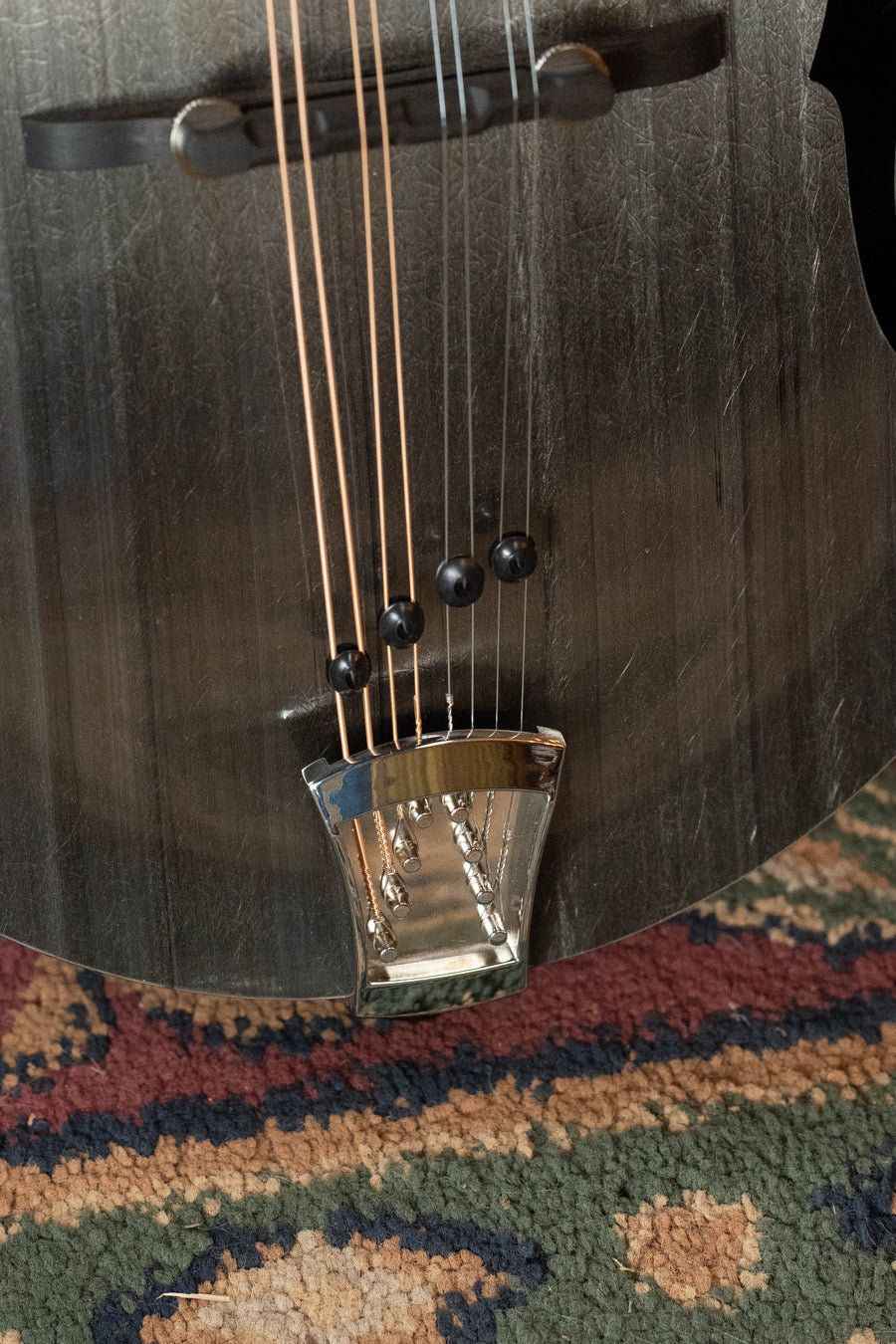Close-up of the KLOS Guitars CT_MA_DA Full Carbon Timber A-Style Acoustic Mandolin #2680, highlighting its metal tailpiece and strings as it rests on a patterned red, green, and beige carpet.