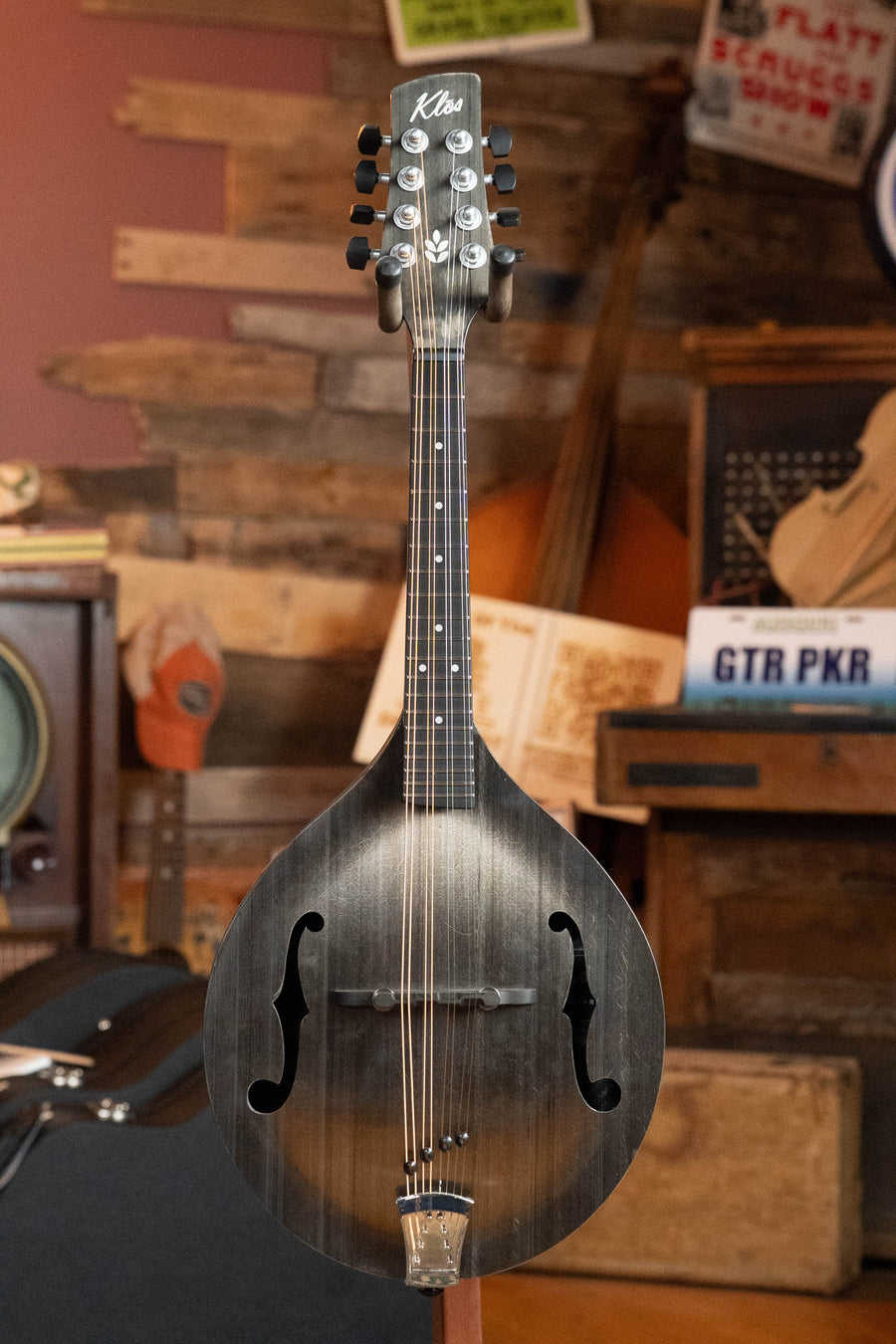 The KLOS Guitars CT_MA_DA Full Carbon Timber A-Style Acoustic Mandolin #2680 stands upright in a rustic room with wooden walls, vintage signs, and musical instruments in the background.