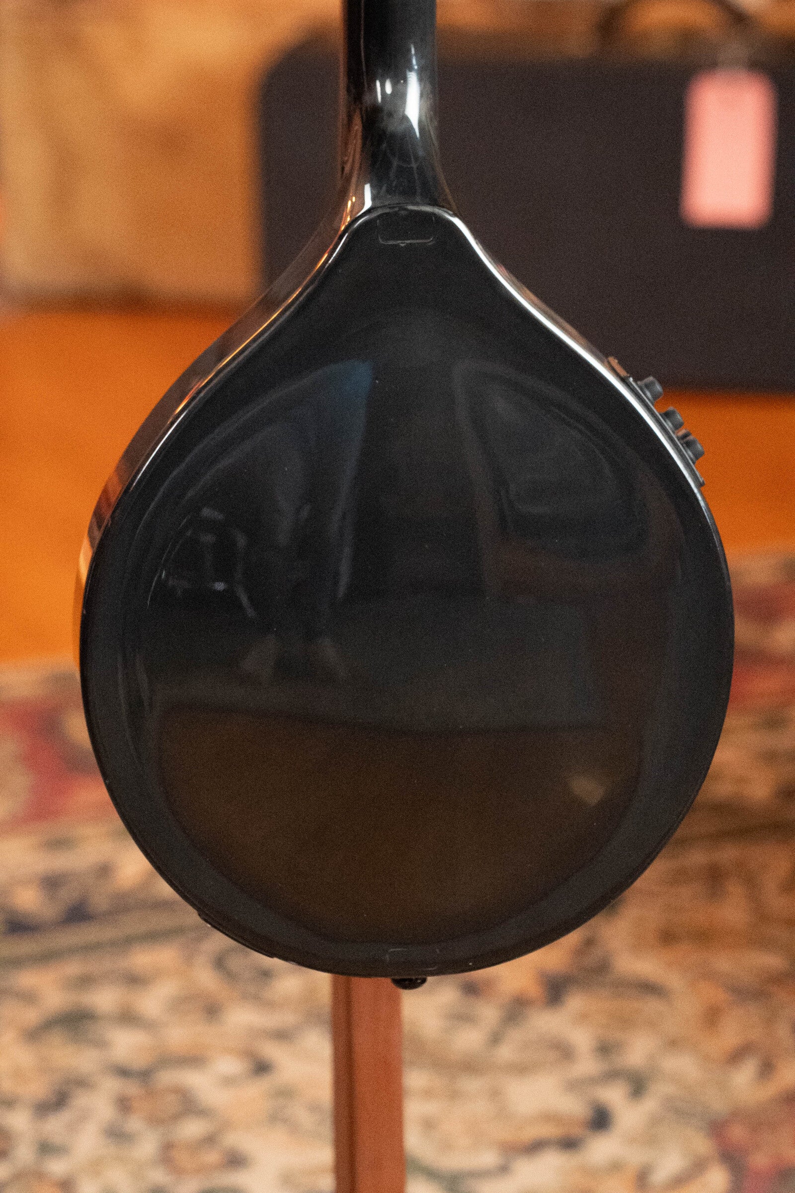 Close-up of the back of a KLOS Guitars CT_MA_DAE_EQ Full Carbon Unidirectional Black A-Style Mandolin with KLOS EQ Pickup (#4889), resting on a stand against a blurred patterned rug and wooden floor.