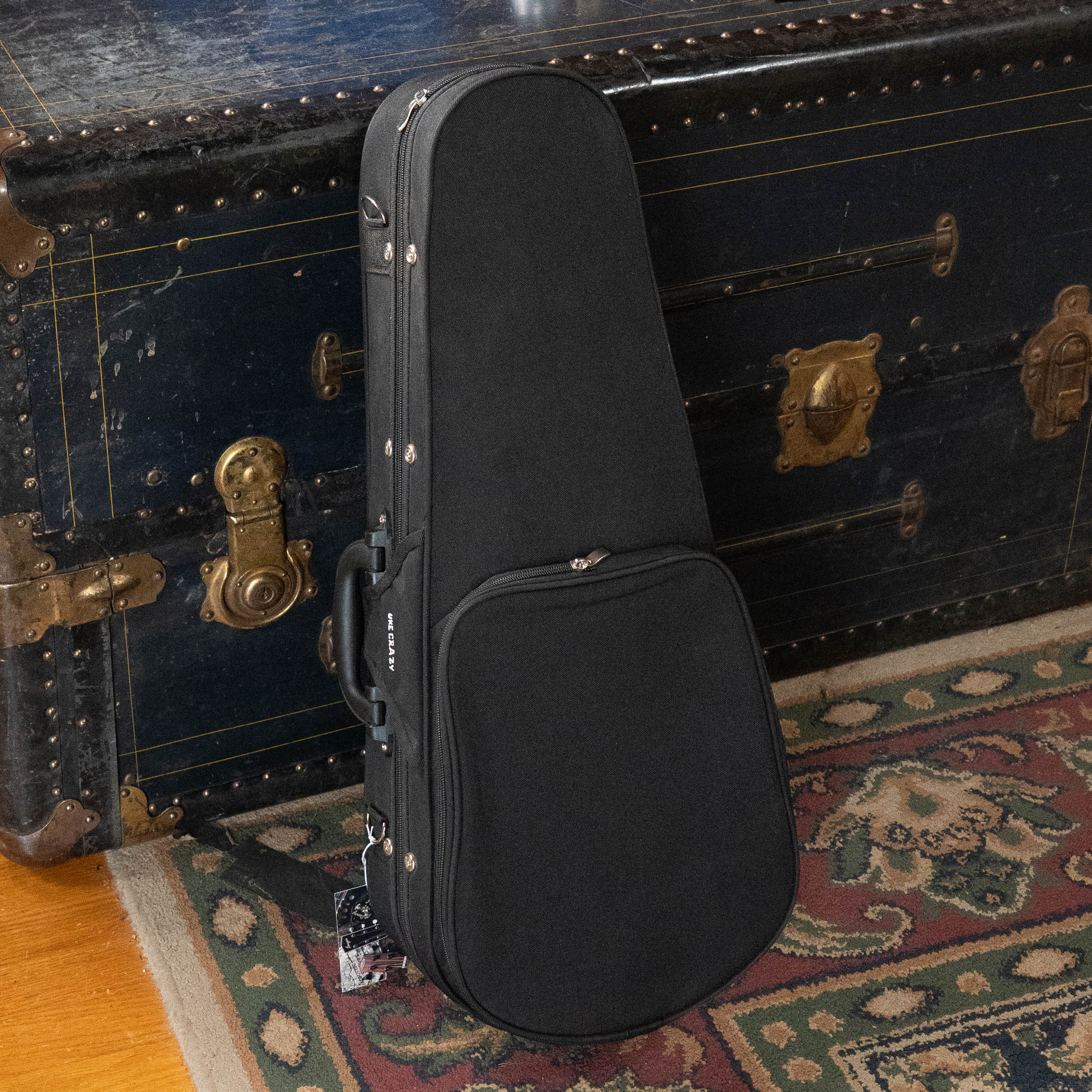The Kala UC-C Foam Concert Ukulele Hardcase by Kala rests against a vintage dark trunk with brass hardware, beside a patterned rug on a wooden floor.